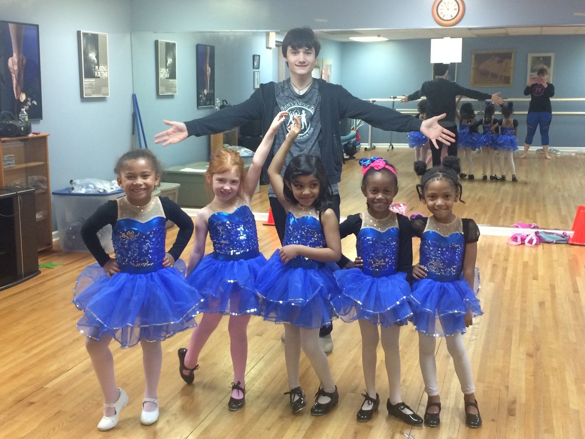 Five young dancers in blue tutus pose with a smiling instructor in a studio mirror room.