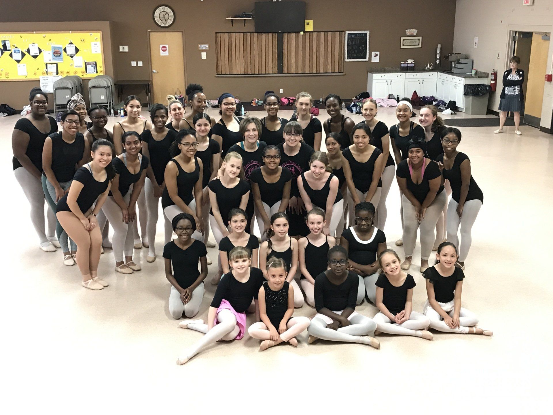 Large group of dancers posing in a studio, with a yellow wall, mirror, and ballet barre in the background.
