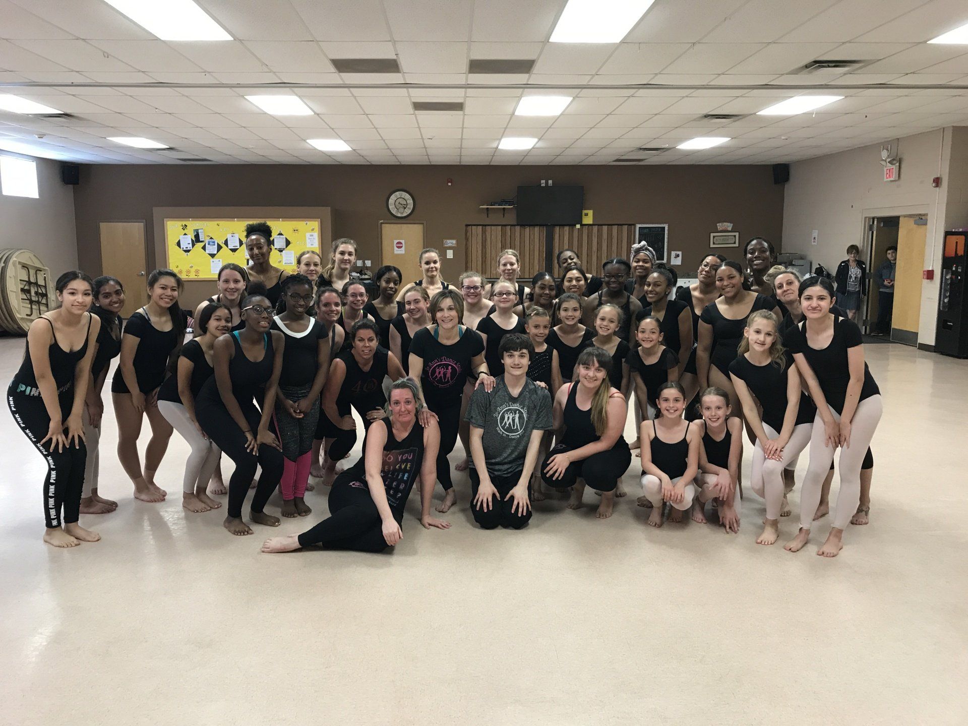 Large dance group posing in a studio, with dancers in black and light outfits lined up for a team photo.