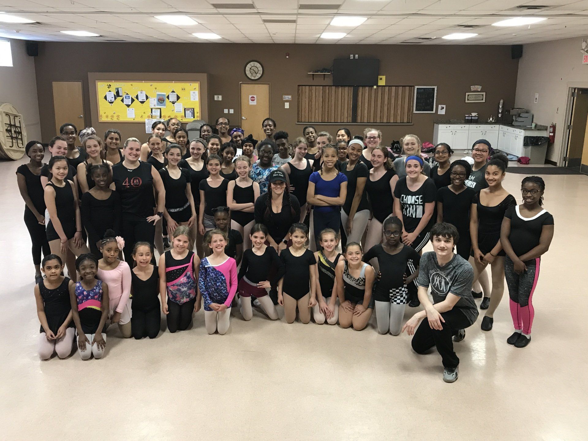 Large group posing in a dance studio, many in black leotards and pink tights, smiling at the camera.