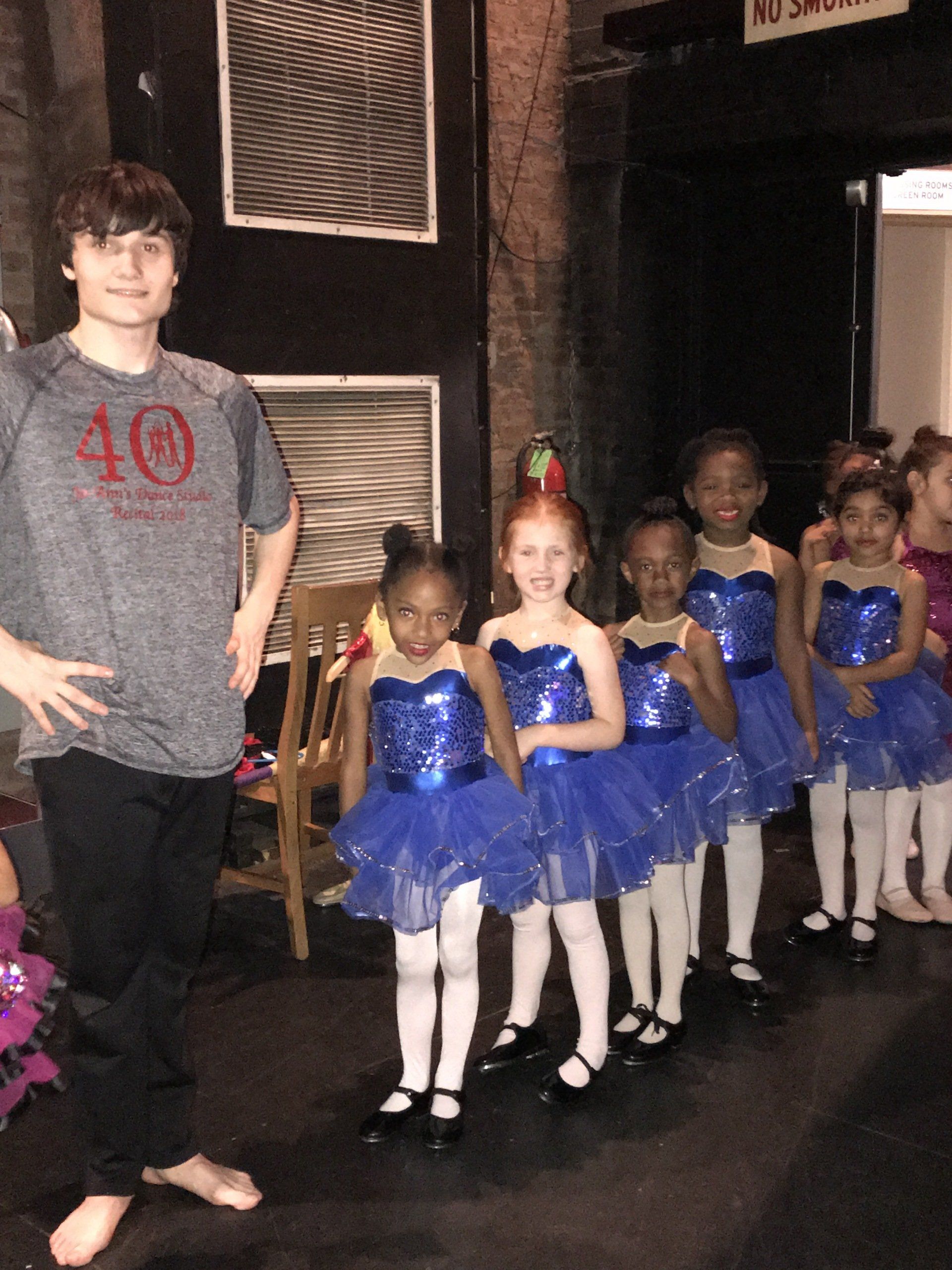 Young dance instructor beside a line of children in blue ballet costumes backstage