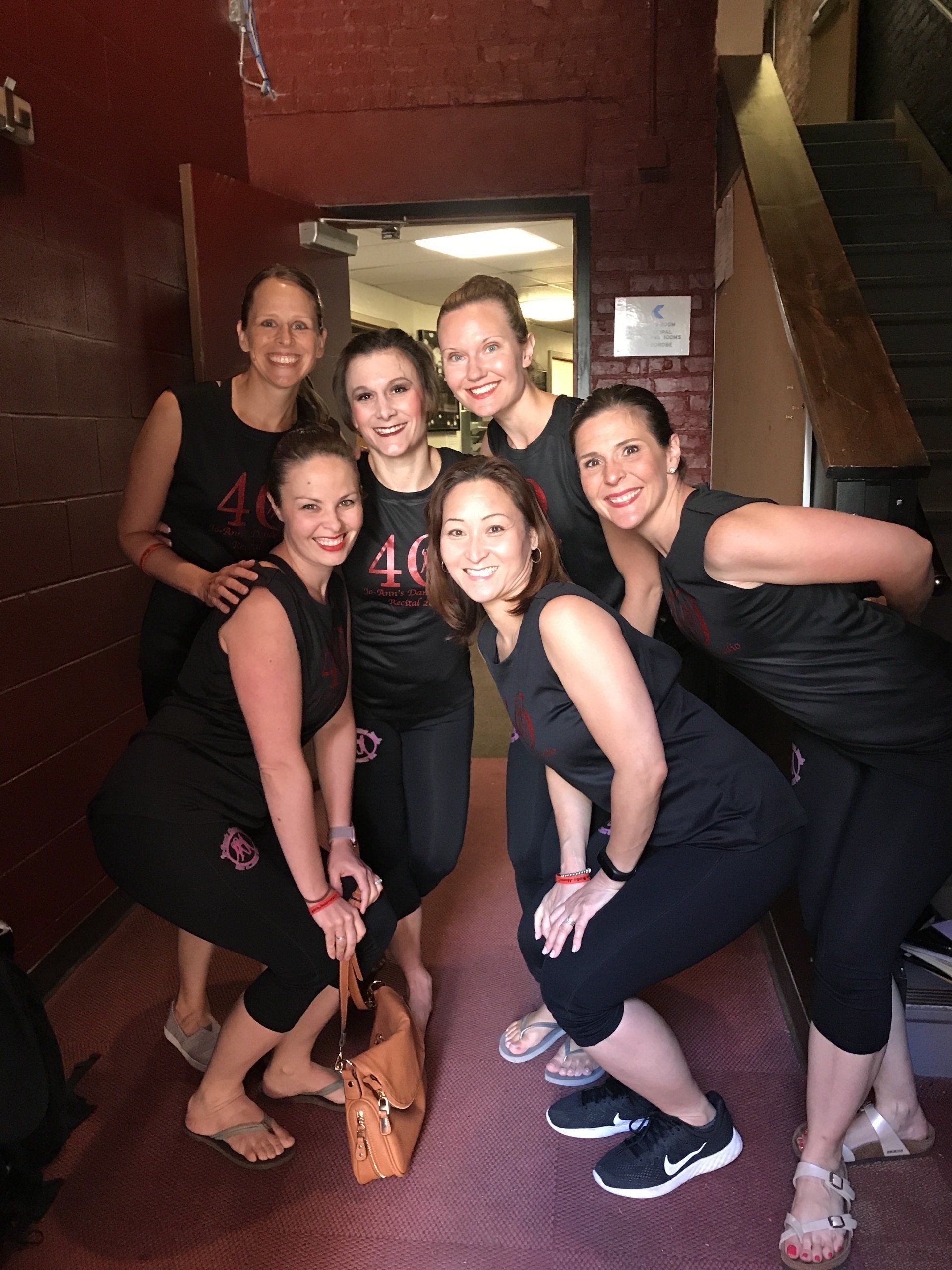 Six women in black outfits posing and smiling together indoors on a wooden floor.