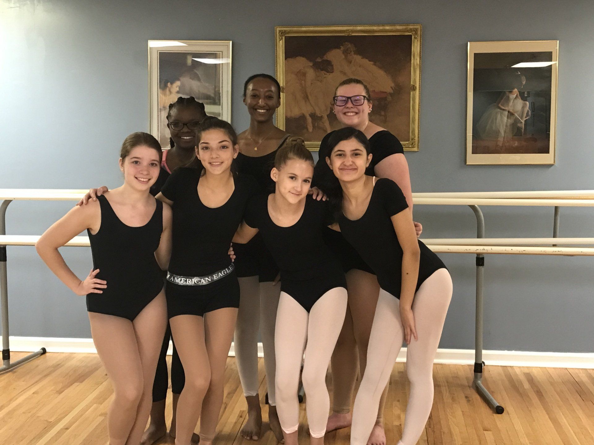 Five dancers in black leotards posing together in a ballet studio with mirrors and barres