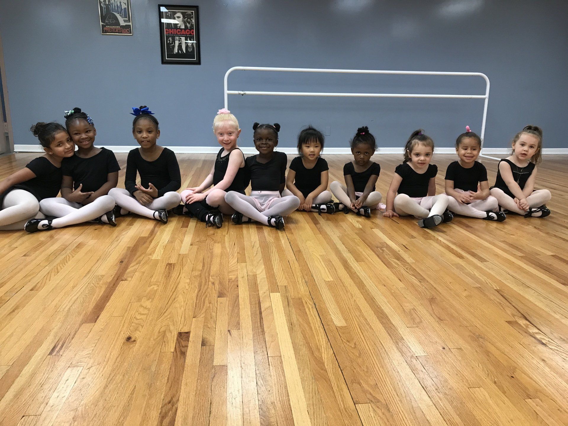 Children in black dance outfits sitting in a row on a wooden studio floor, facing forward.