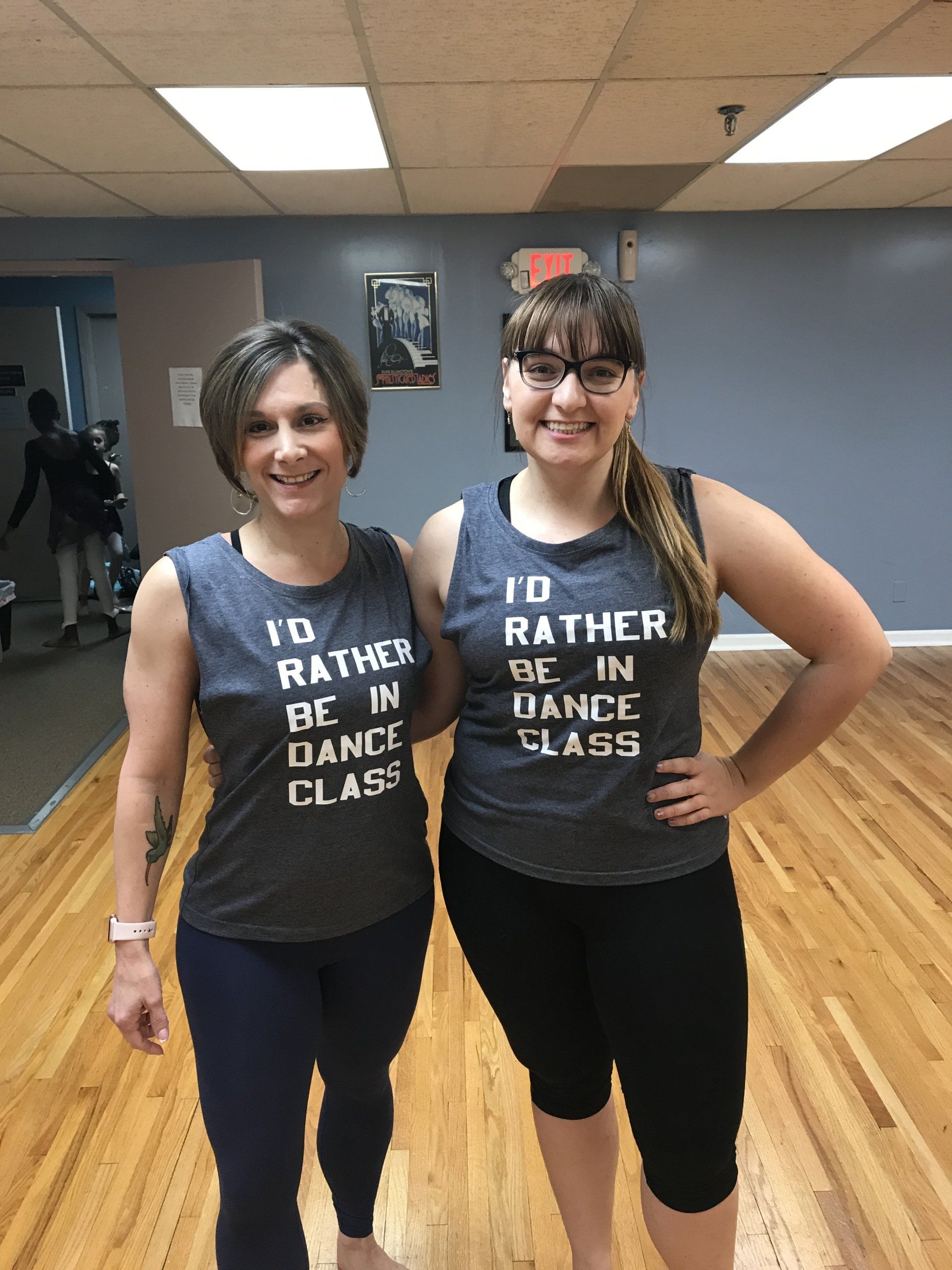 Two people standing in a dance studio, smiling and wearing matching gray workout shirts.