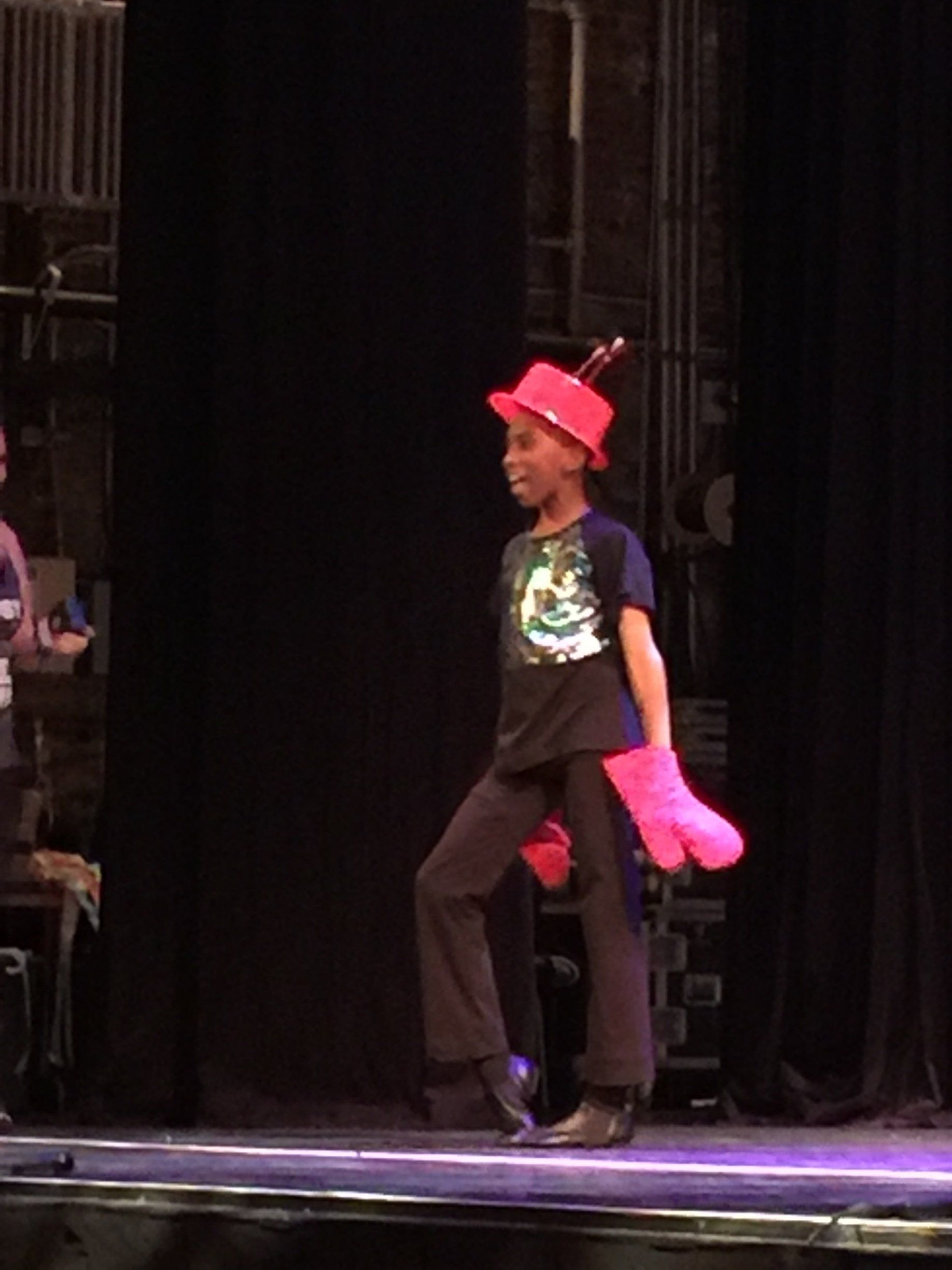 Person on a stage wearing a red hat and bright pink gloves, mid-step in a playful pose.