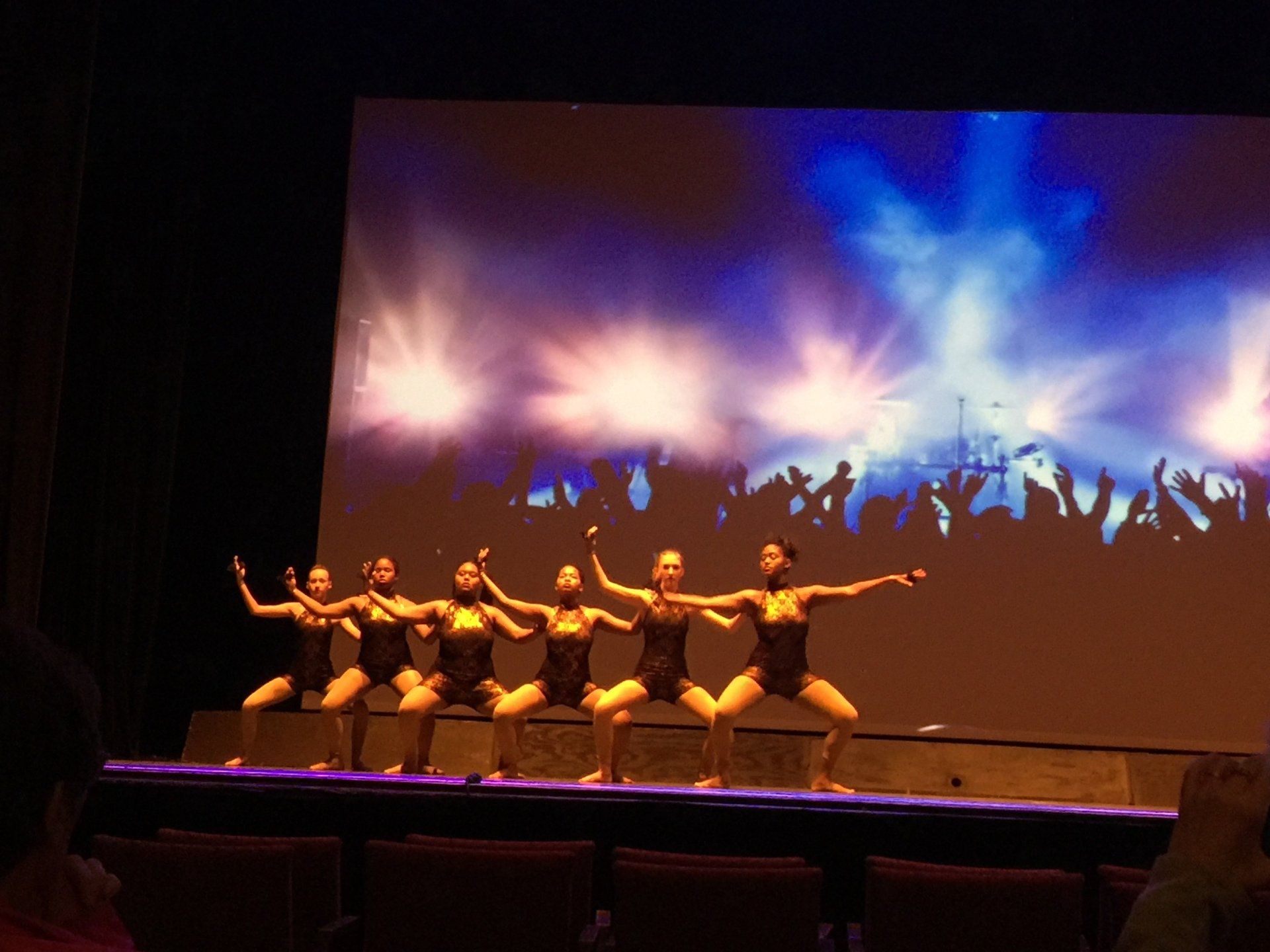 Six dancers pose onstage in synchronized ballet under colorful blue and pink lights