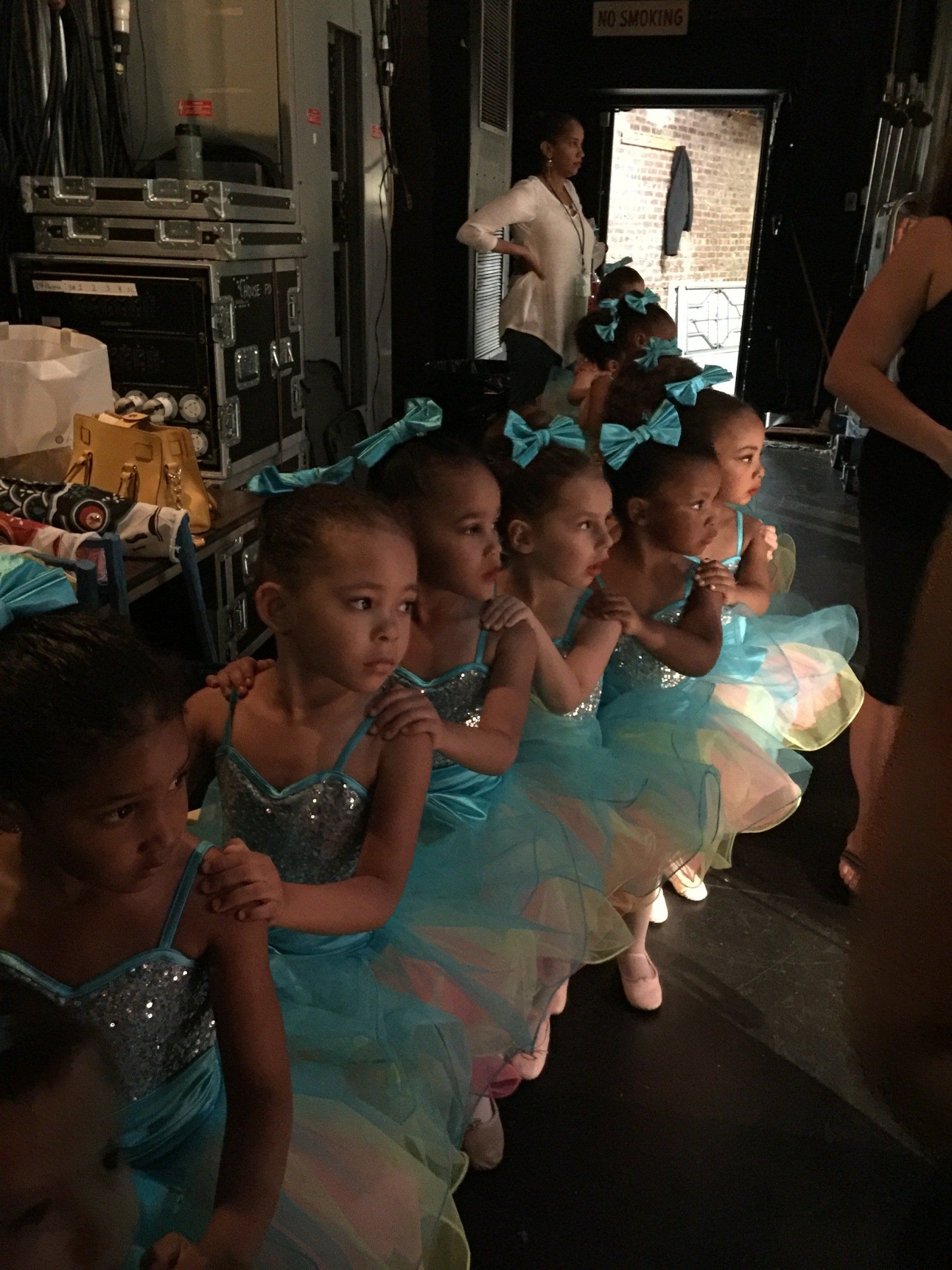 Children in turquoise costumes sit backstage, waiting for a dance performance.