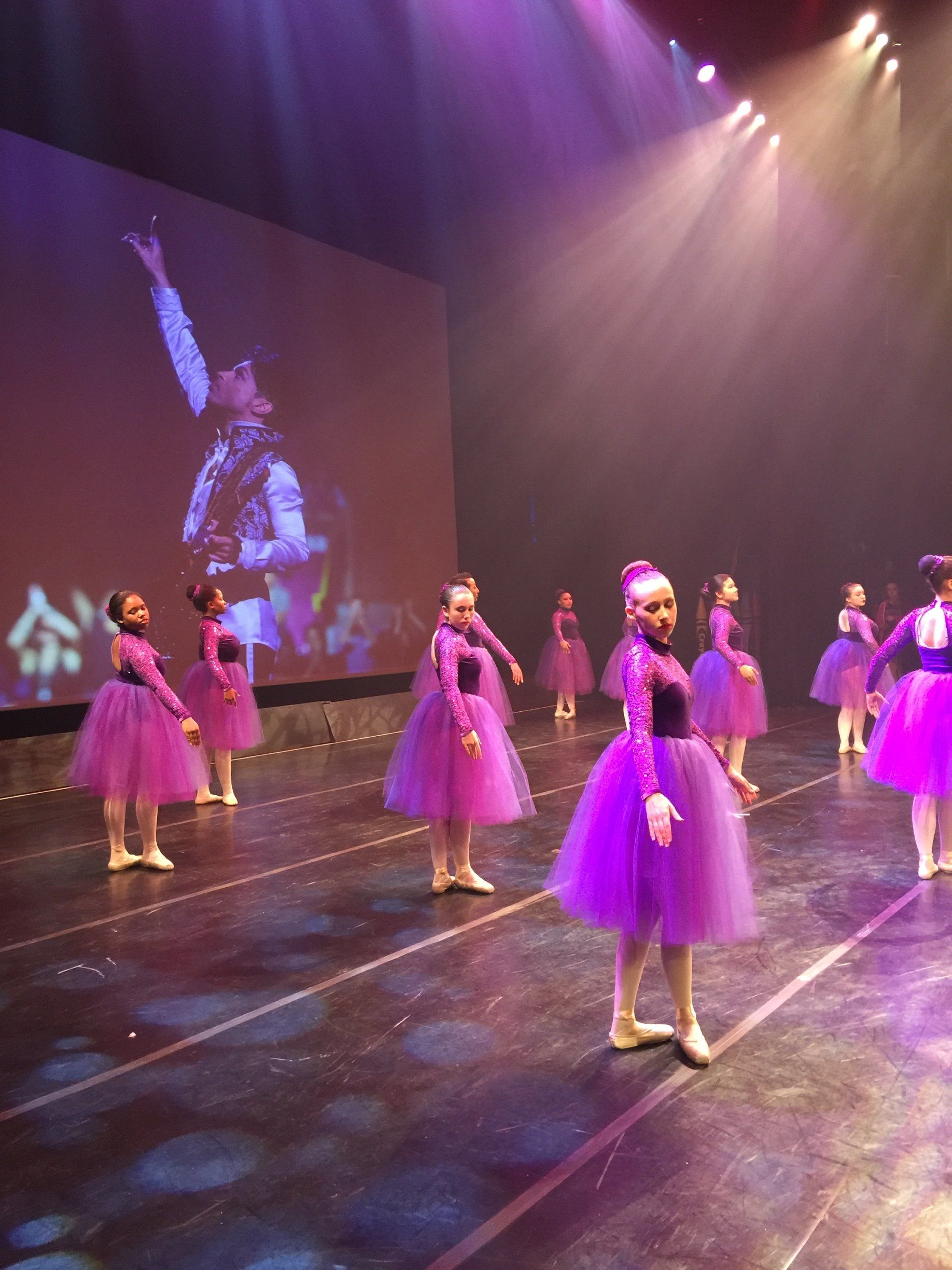 Children in purple tutus performing on a stage under pink and blue lights
