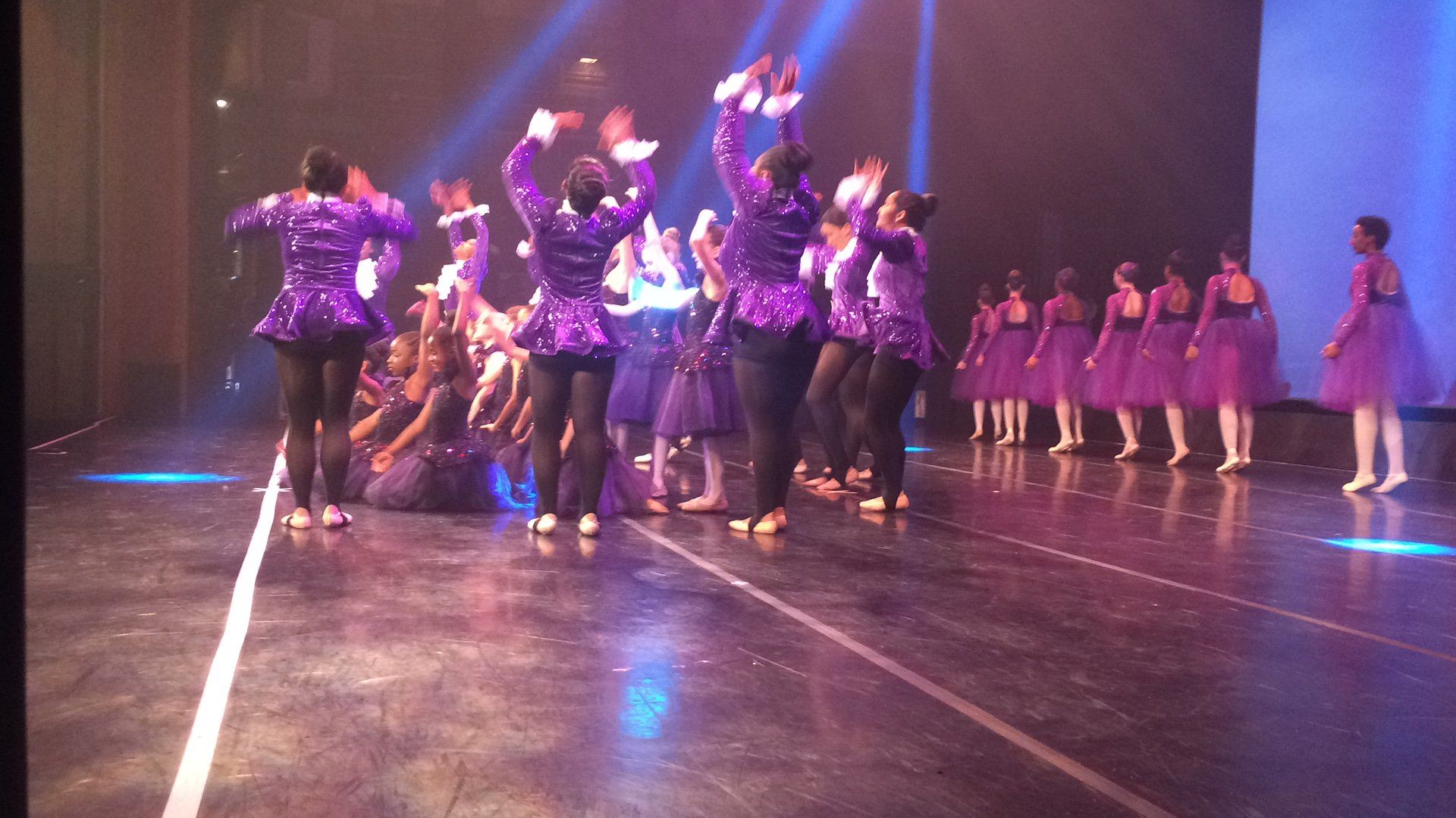 Stage dance troupe in purple costumes performing under purple lights, with dancers lined up in the background
