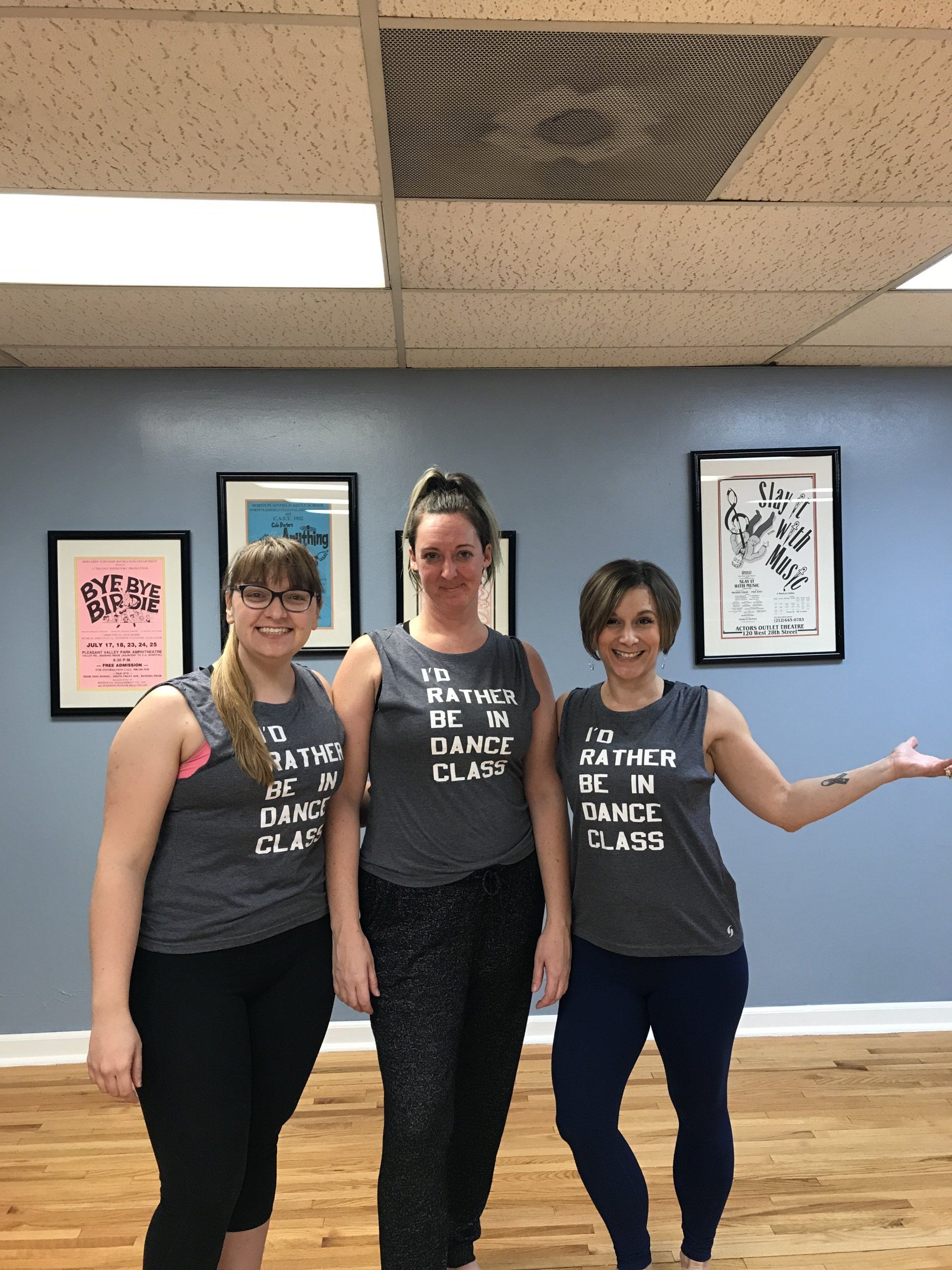 Three people posing indoors in matching black tank tops in front of framed wall art and a blue wall.