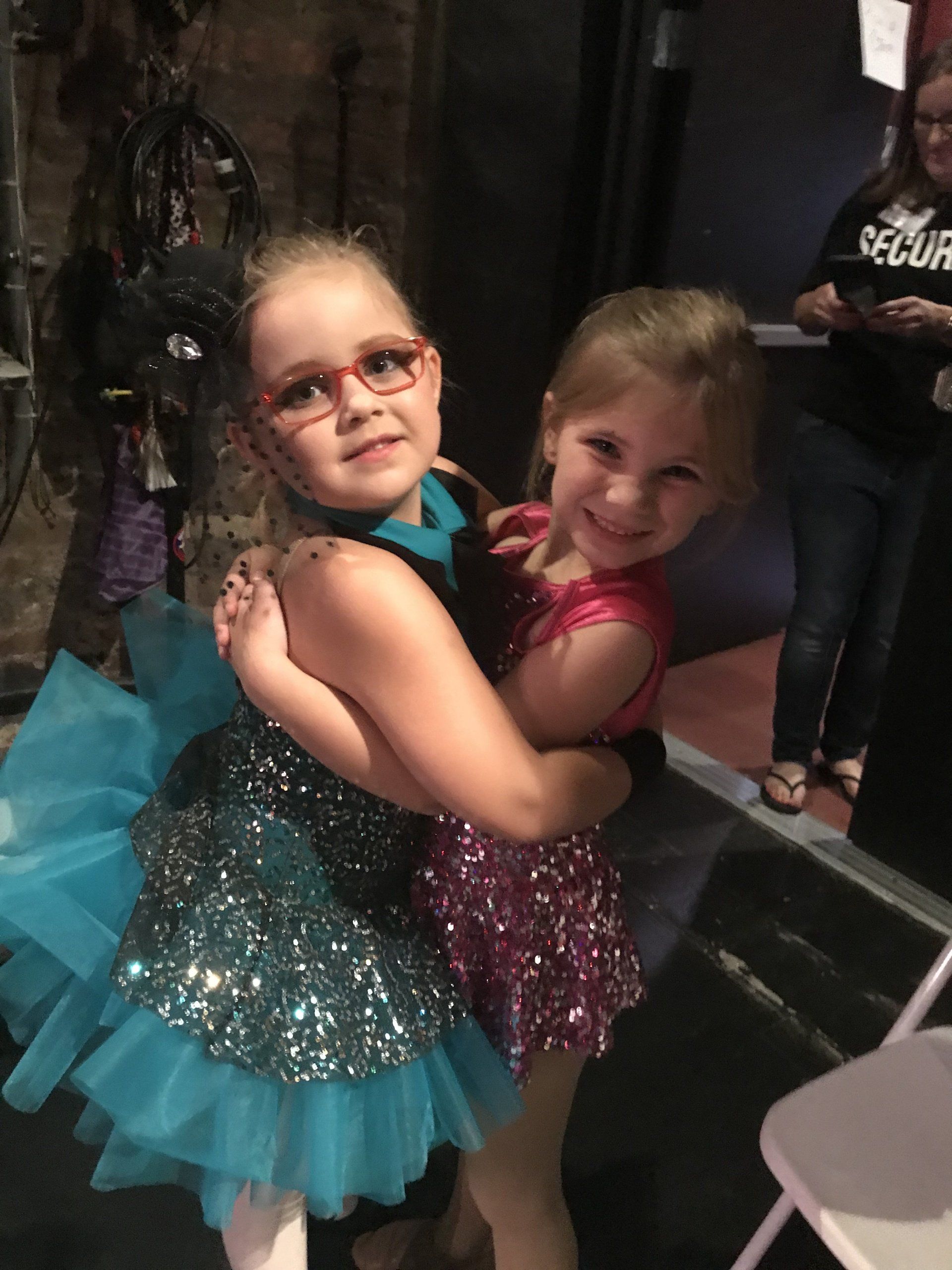 Two children in sparkly dresses hugging and smiling at an indoor event.