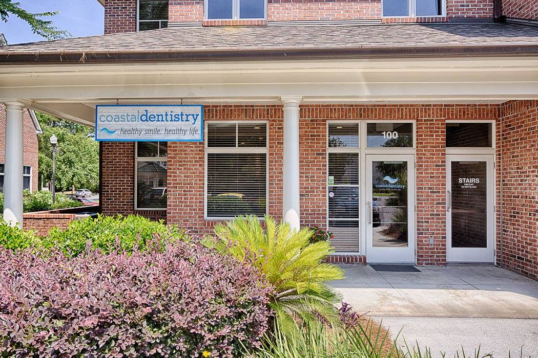 Brick building entrance with glass doors, blue sign, and pink shrubs in front