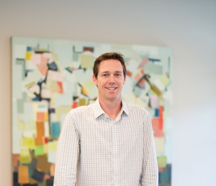 Man standing indoors in front of a colorful abstract painting, wearing a light checkered shirt, smiling.
