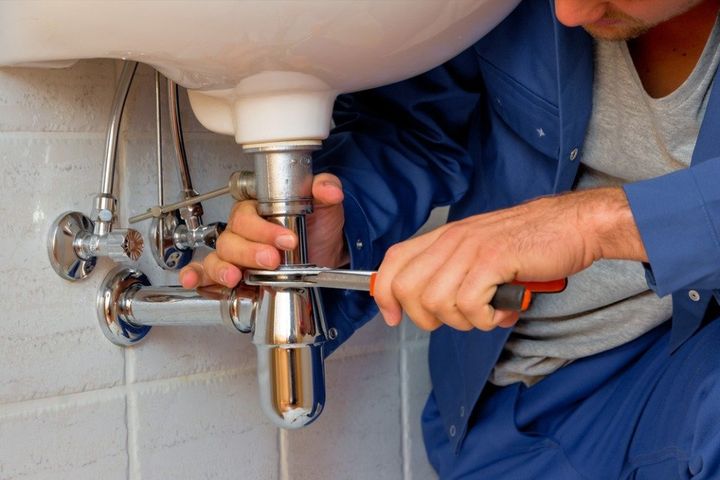 Un idraulico stringe i tubi sotto un lavandino con una chiave inglese in un bagno piastrellato.