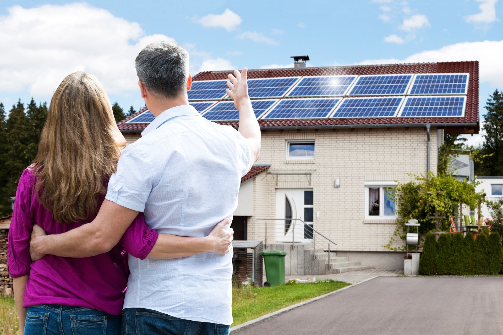 Ein Paar betrachtet ein Haus mit Solarpaneelen auf dem Dach. Der Mann zeigt nach oben; es ist ein sonniger Tag.