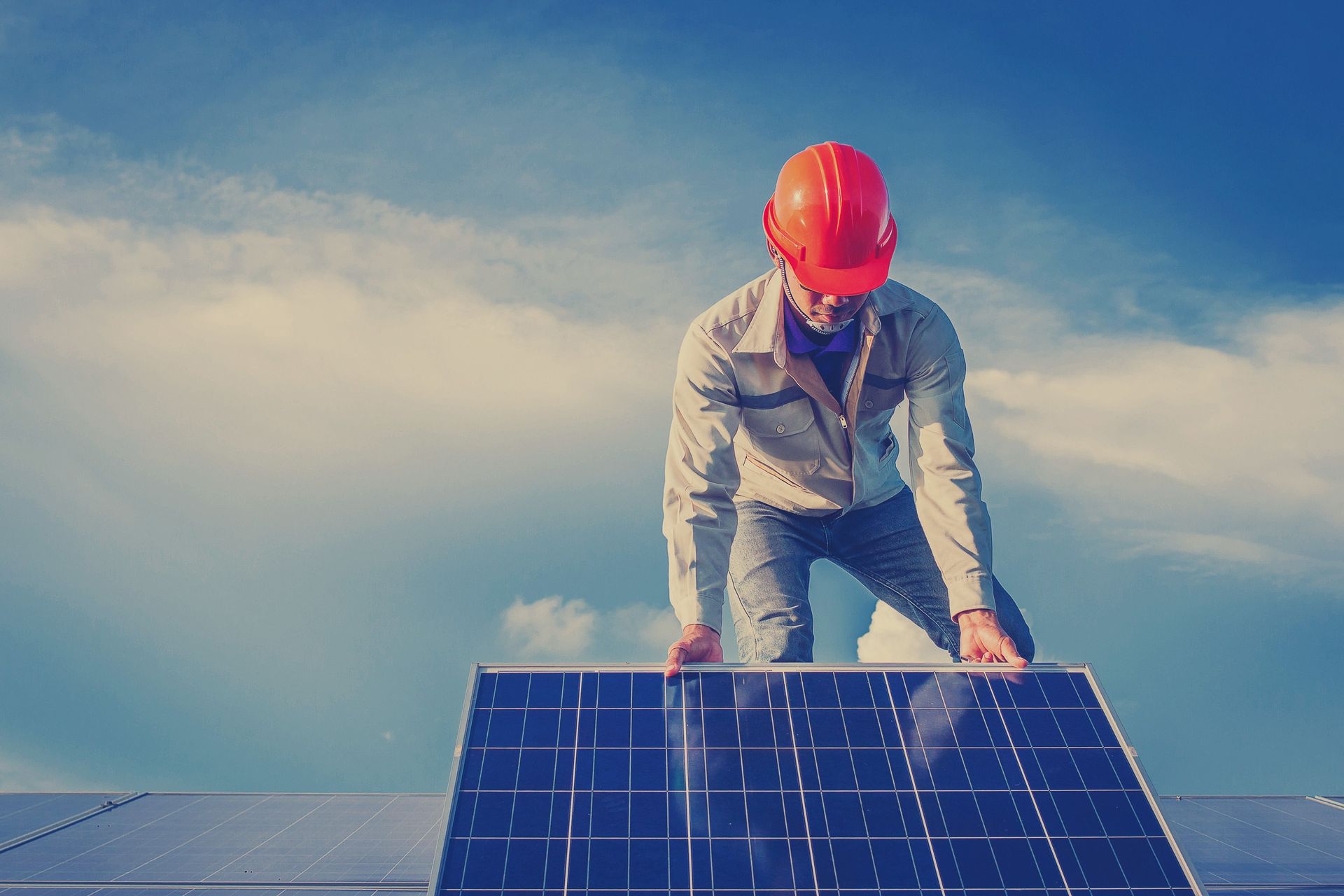 Ein Arbeiter mit orangefarbenem Schutzhelm installiert unter blauem Himmel ein Solarpanel auf einem Dach.