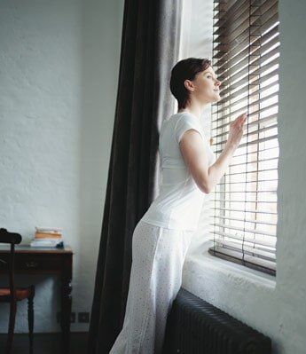 Woman looking out a window with slatted blinds, leaning forward. She wears white and looks pensive.