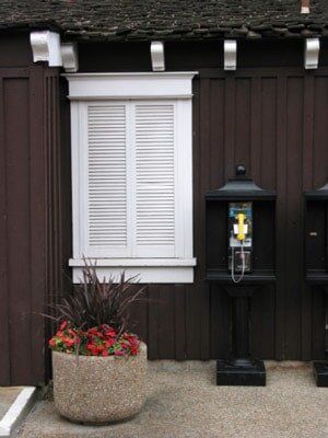 Brown building facade with white shuttered window, black payphone, and flower pot.