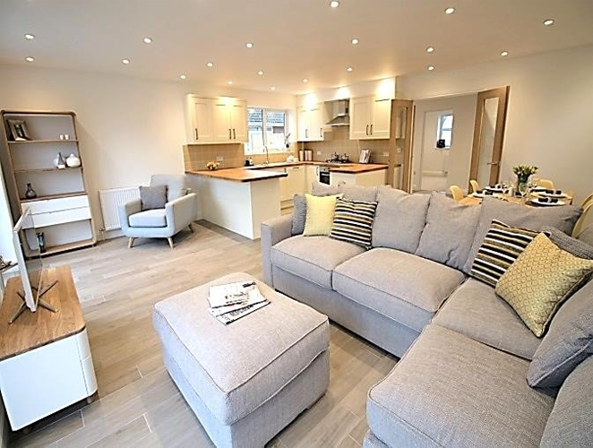 Bright open-plan living room with gray sectional sofa, ottoman, and kitchen in the background.