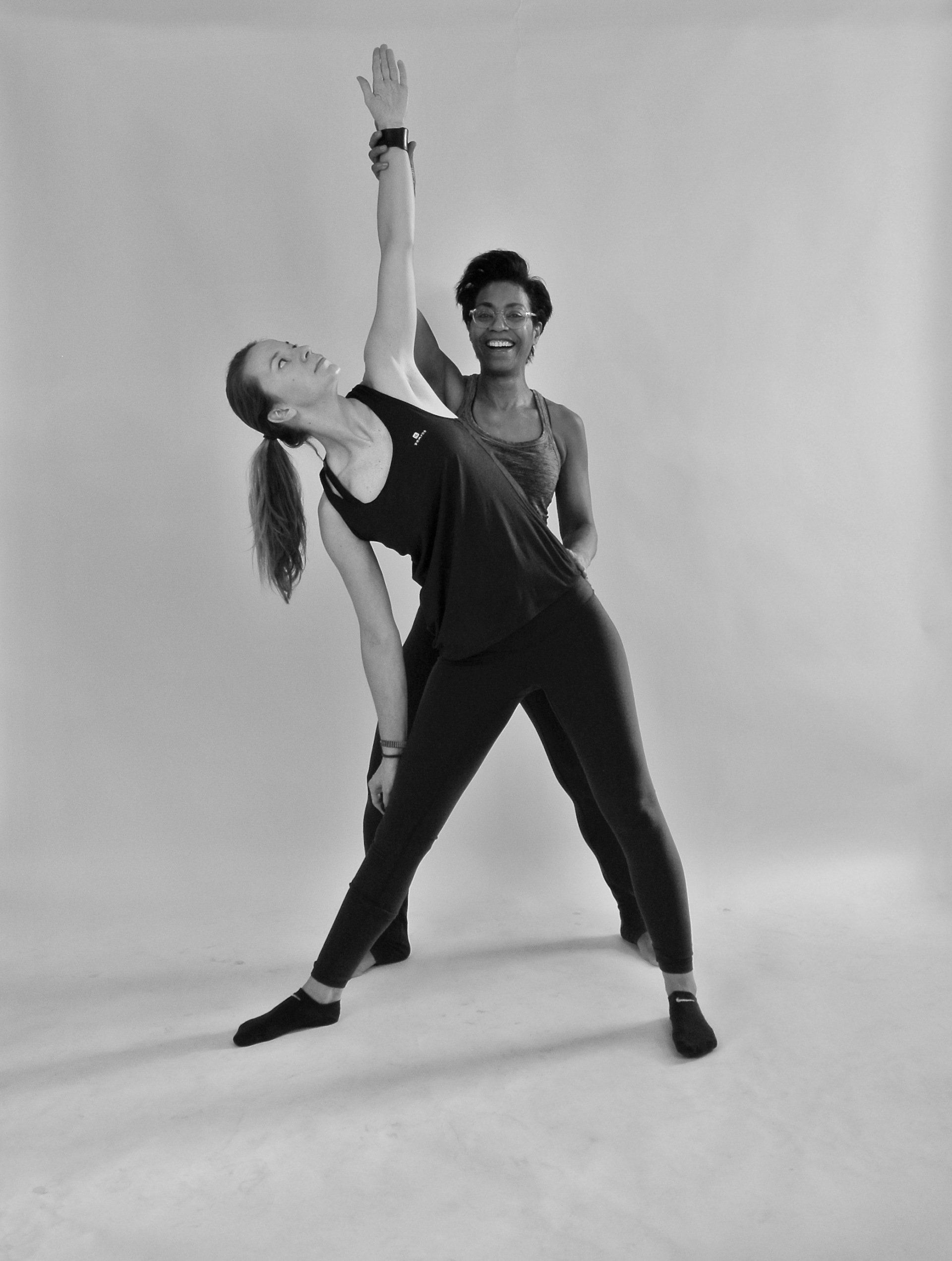 Twee dansers poseren in zwarte outfits tegen een effen studioachtergrond, met één arm omhoog en hun lichamen iets uit elkaar.