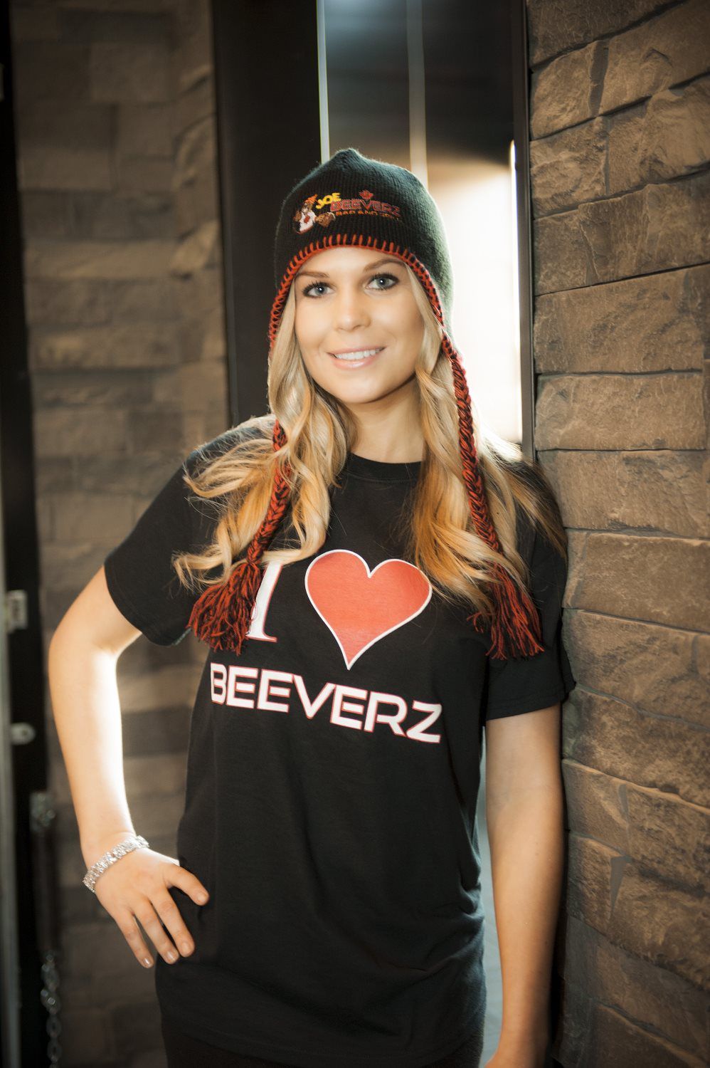 Woman in black “I ♥ BEEVERZ” T-shirt and beanie, smiling and standing by a stone wall