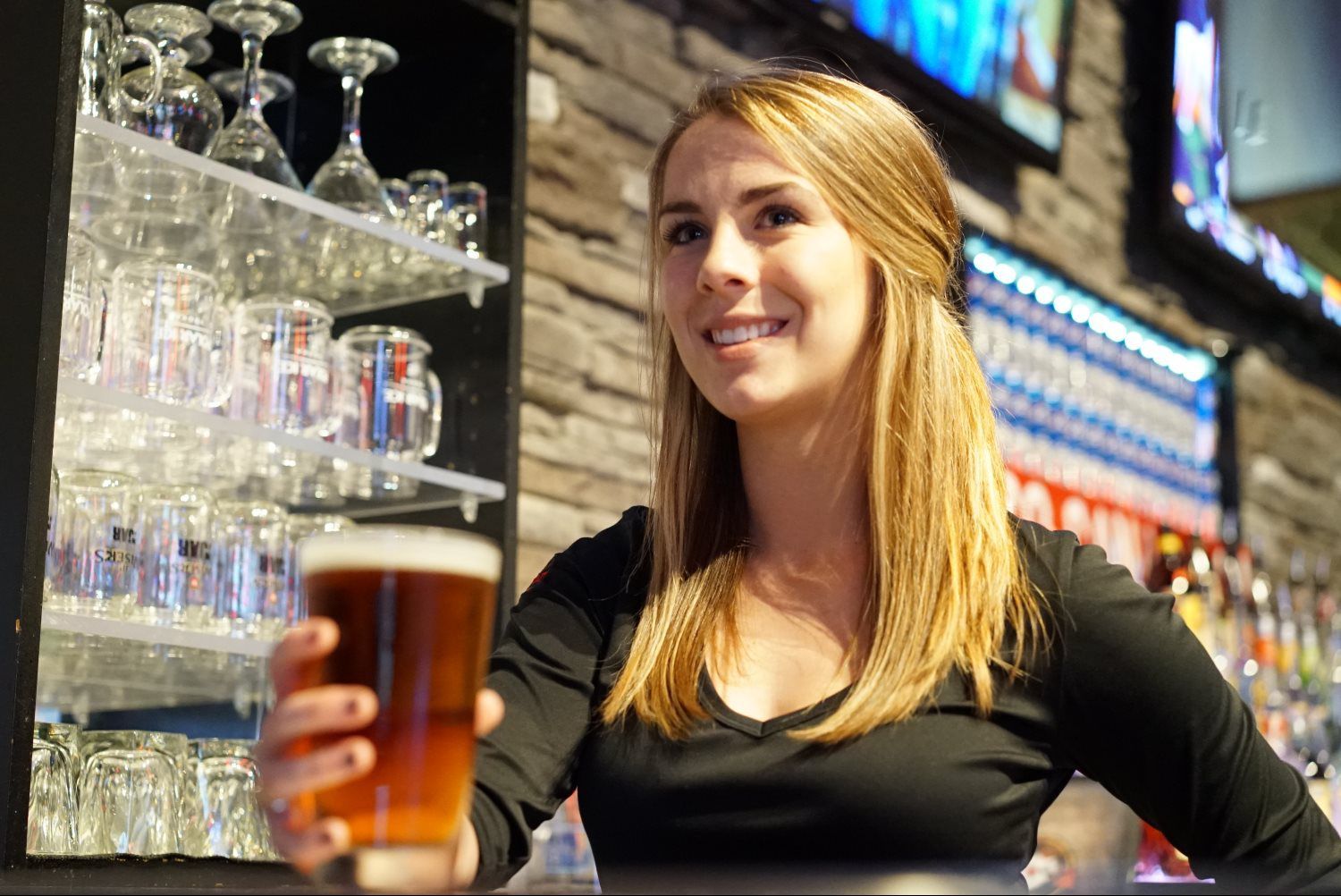 Bartender smiling behind a bar, holding out a pint of beer in a lively pub.