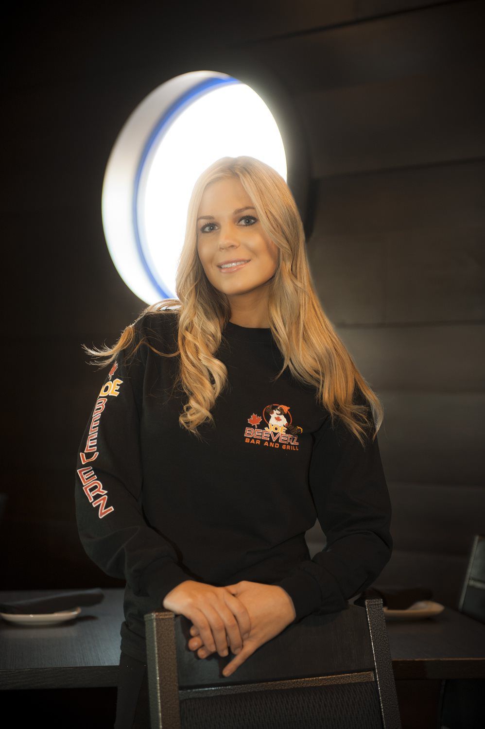 Woman standing indoors beside a glowing circular light, wearing a black top and smiling at the camera.