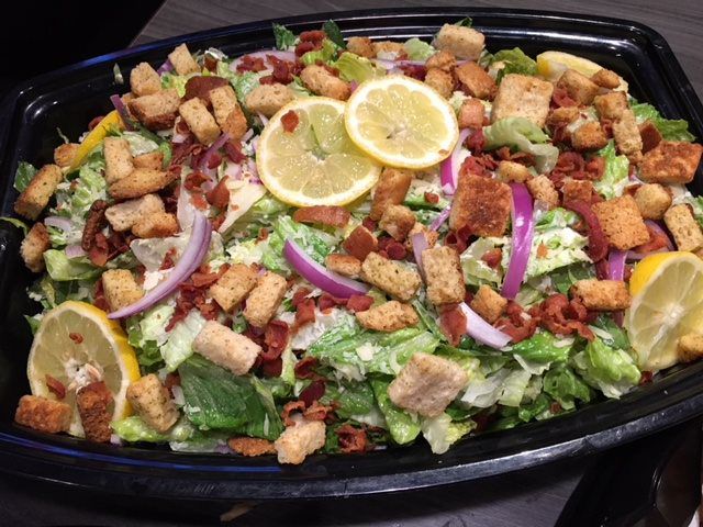 Caesar salad in a black tray with croutons, lettuce, red onion, lemon slices, and dressing