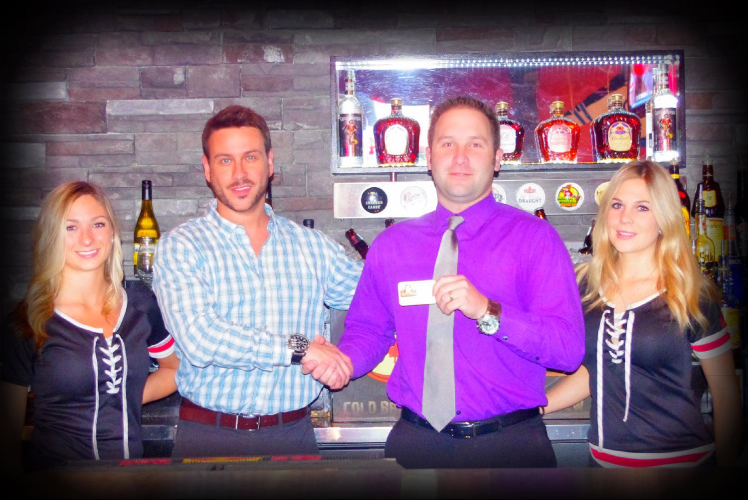 Four people posing in a bar, smiling beside shelves of bottles and award plaques.