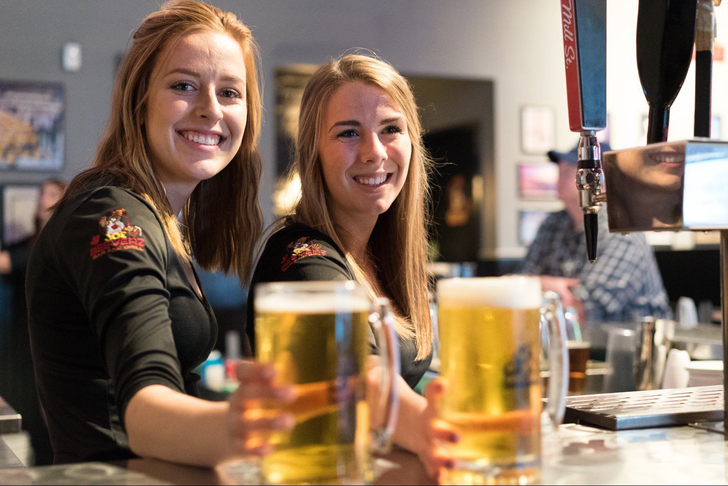 Two smiling bartenders behind a bar with glasses and beer taps in the foreground