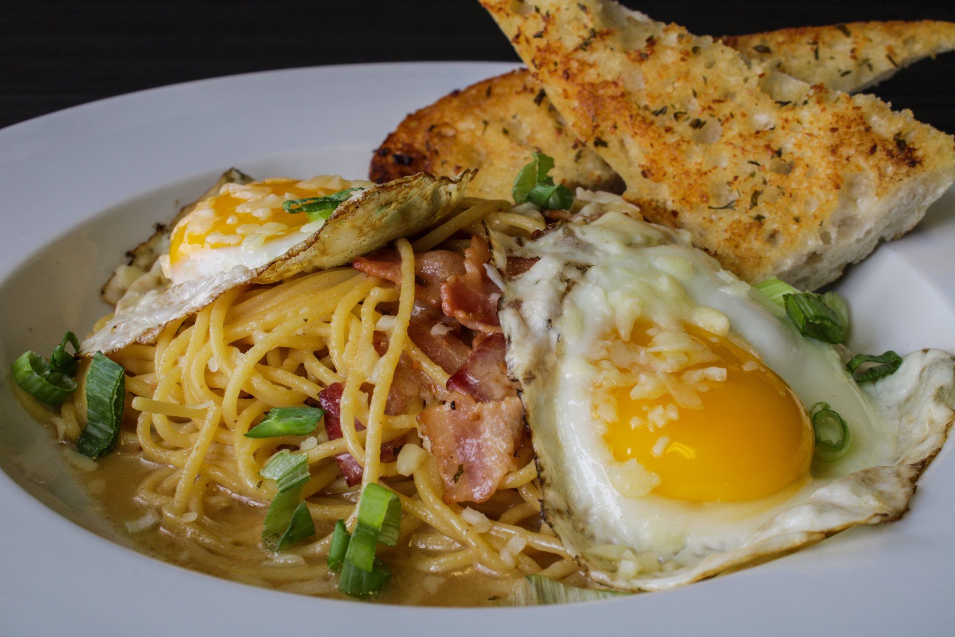 Pasta carbonara with fried eggs, bacon, and garlic bread on a white plate