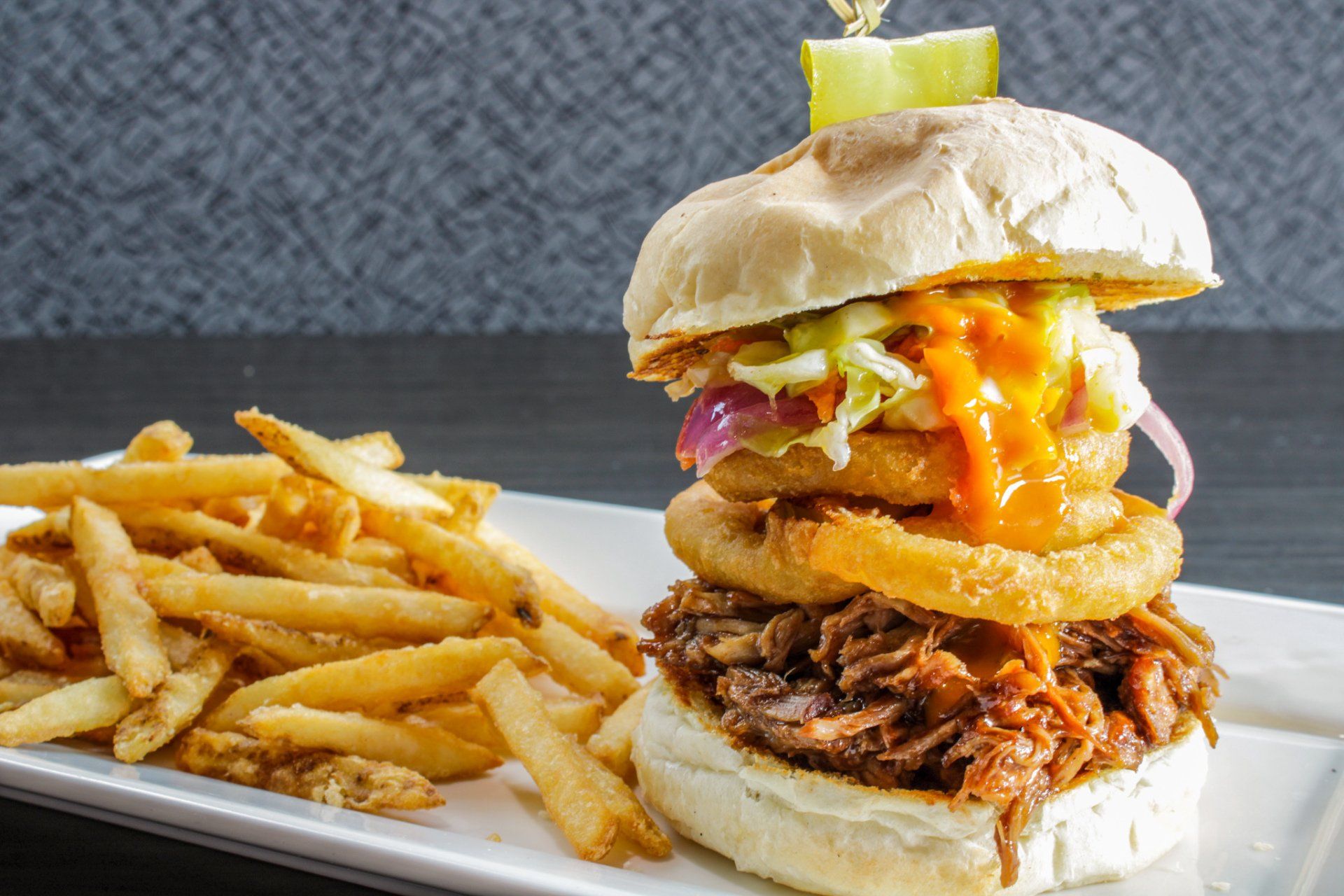 Sandwich stacked with onions, fried rings, pulled pork, and melted cheese beside fries on a plate