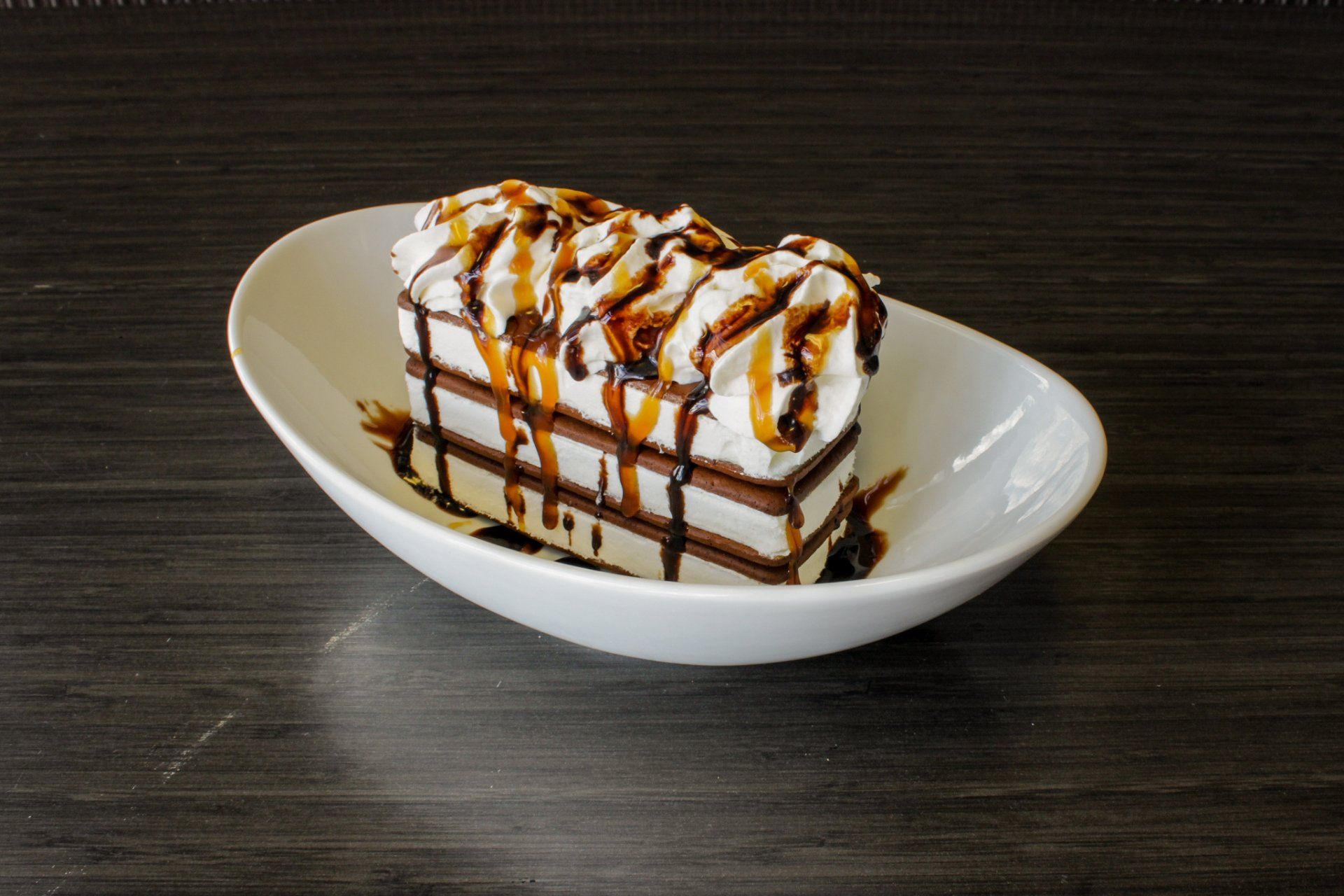 Dessert cake slice with whipped cream and chocolate drizzle in a white bowl on a dark table