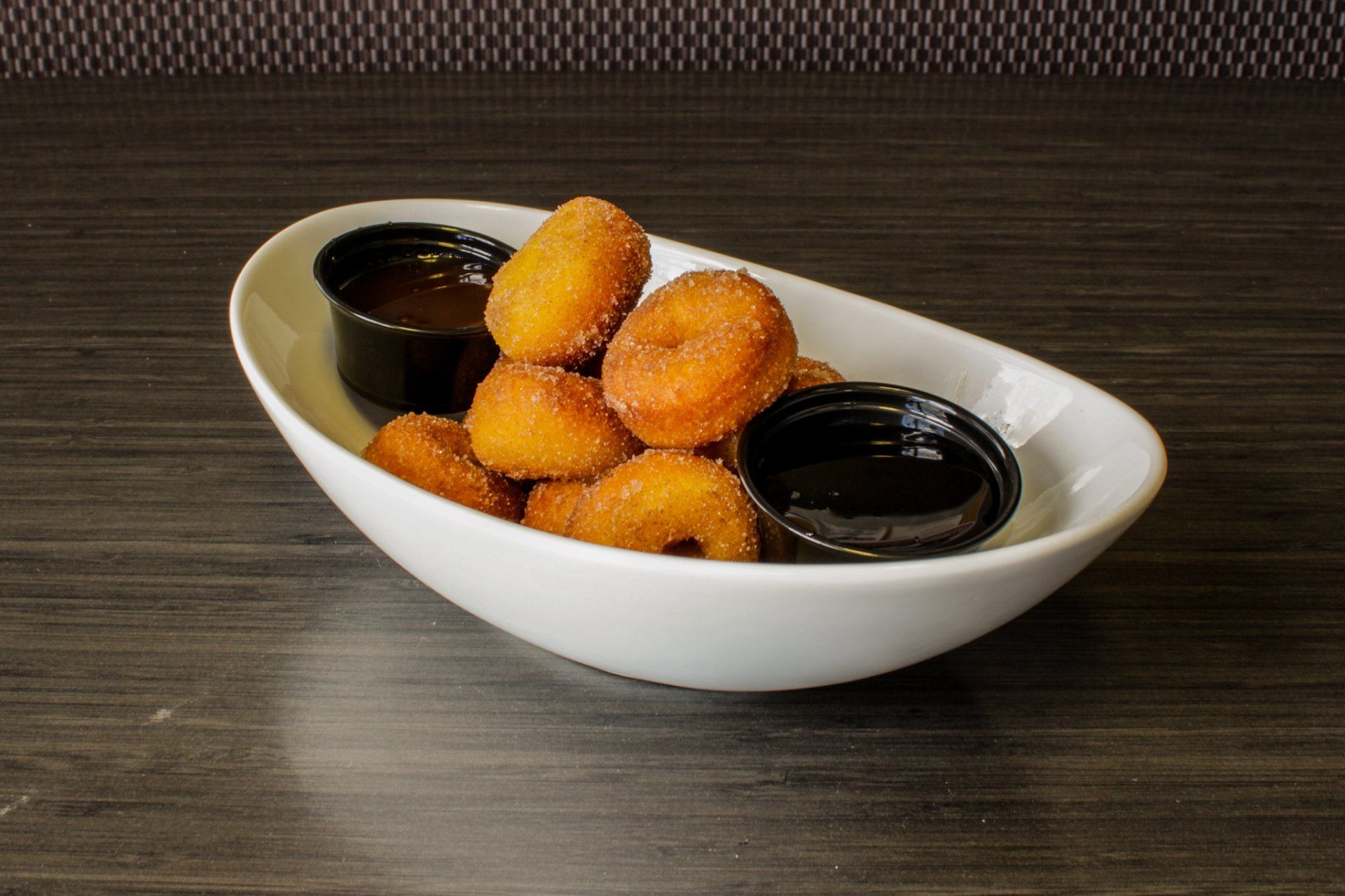 Golden fried bites in a white bowl with two black dipping sauces on a dark table