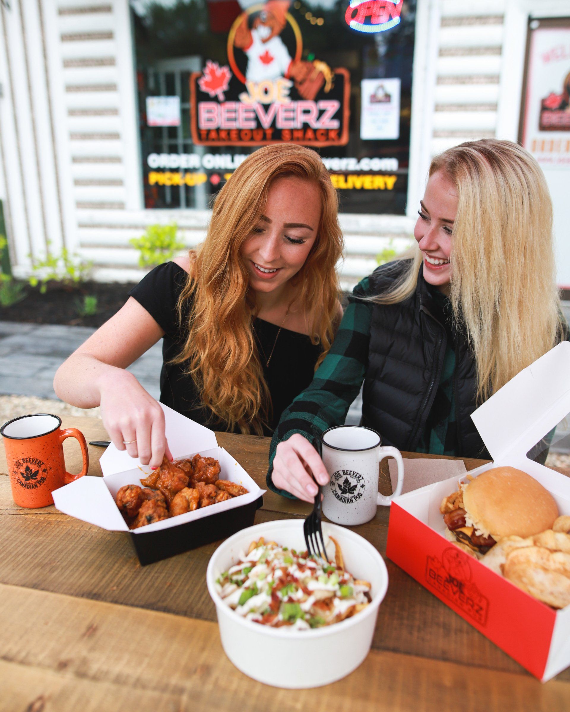 Two people sharing fast food outdoors at a table, smiling over burgers, fries, and drinks.