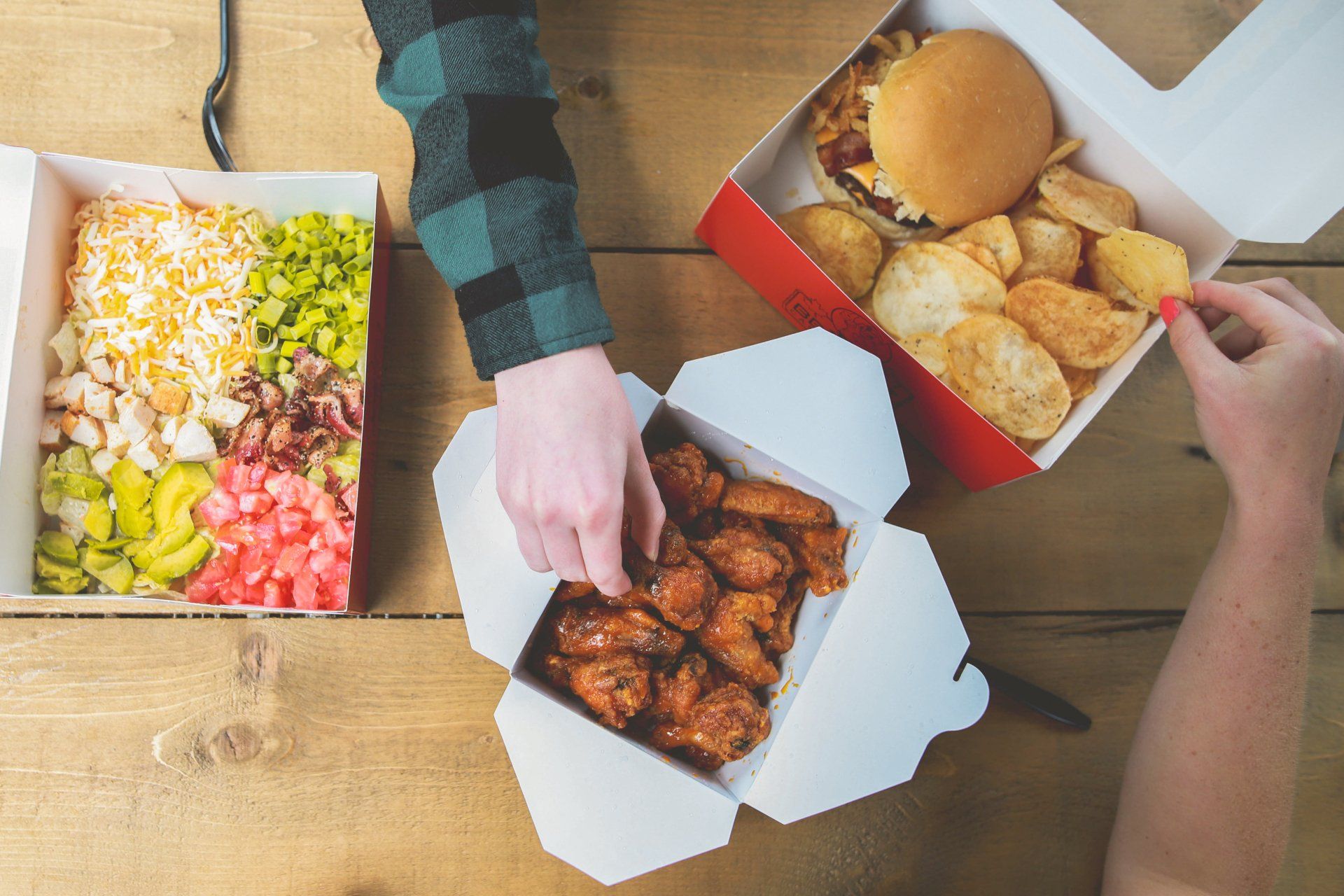 Hands reaching for takeout boxes of pizza, fried chicken, and salad on a wooden table.