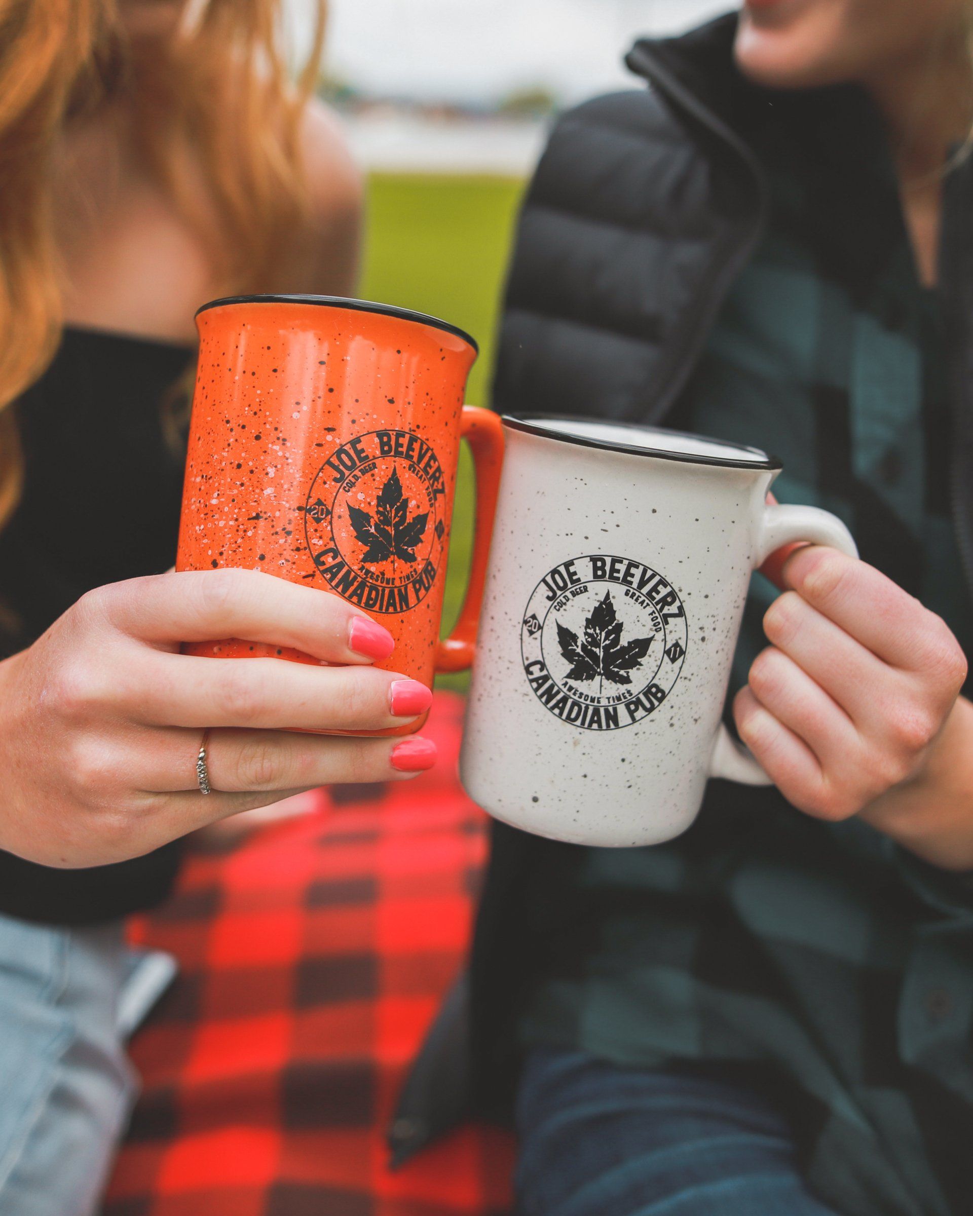 Two people clink orange and white mugs outdoors, one in a red plaid shirt and one in a black jacket.