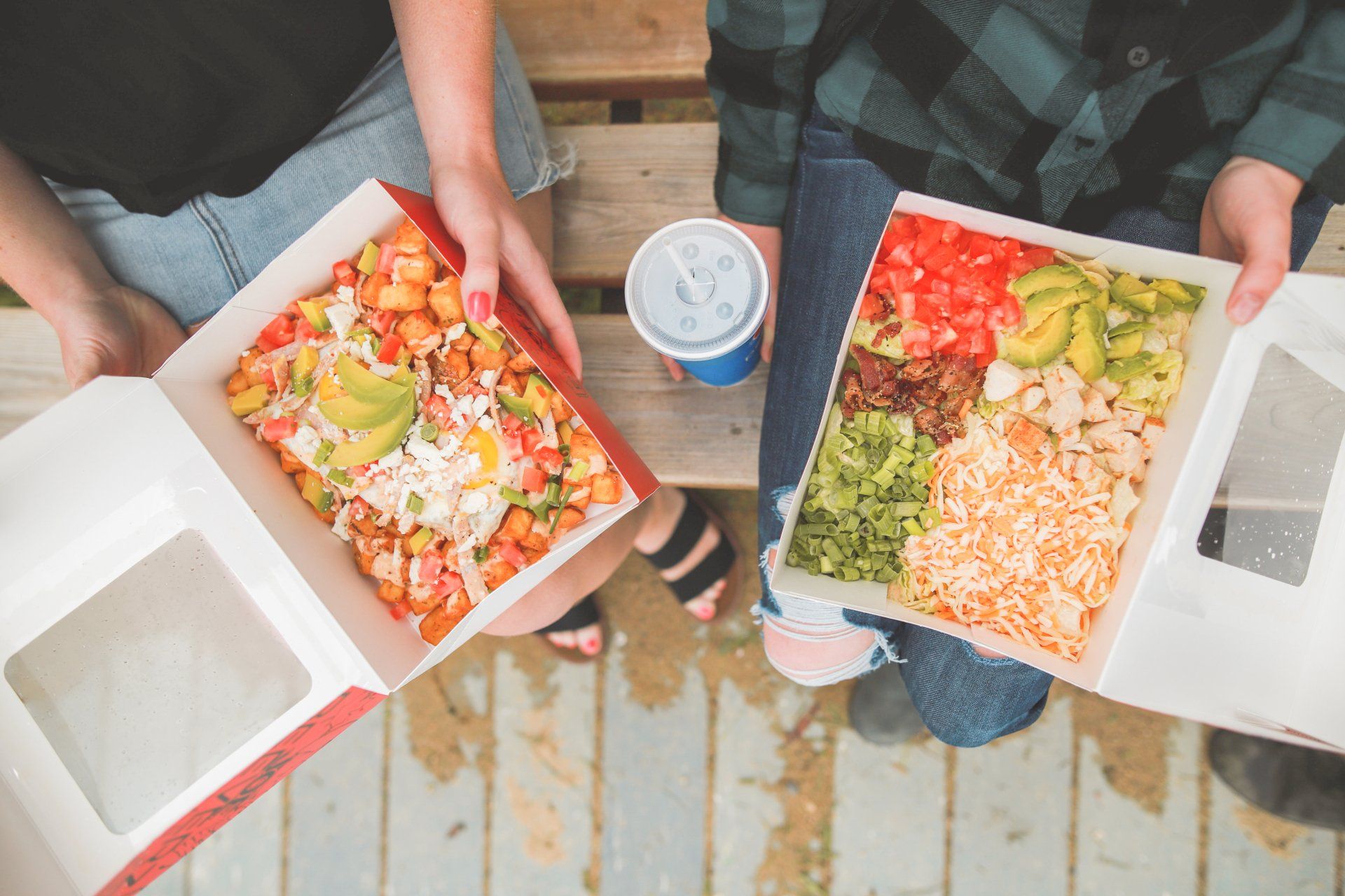 Two people holding open boxes of colorful salad and a drink outdoors on wooden planks