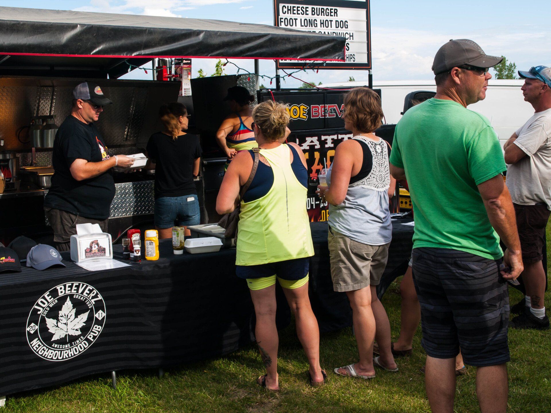 People waiting at an outdoor food booth with a black canopy and a staff member serving drinks.
