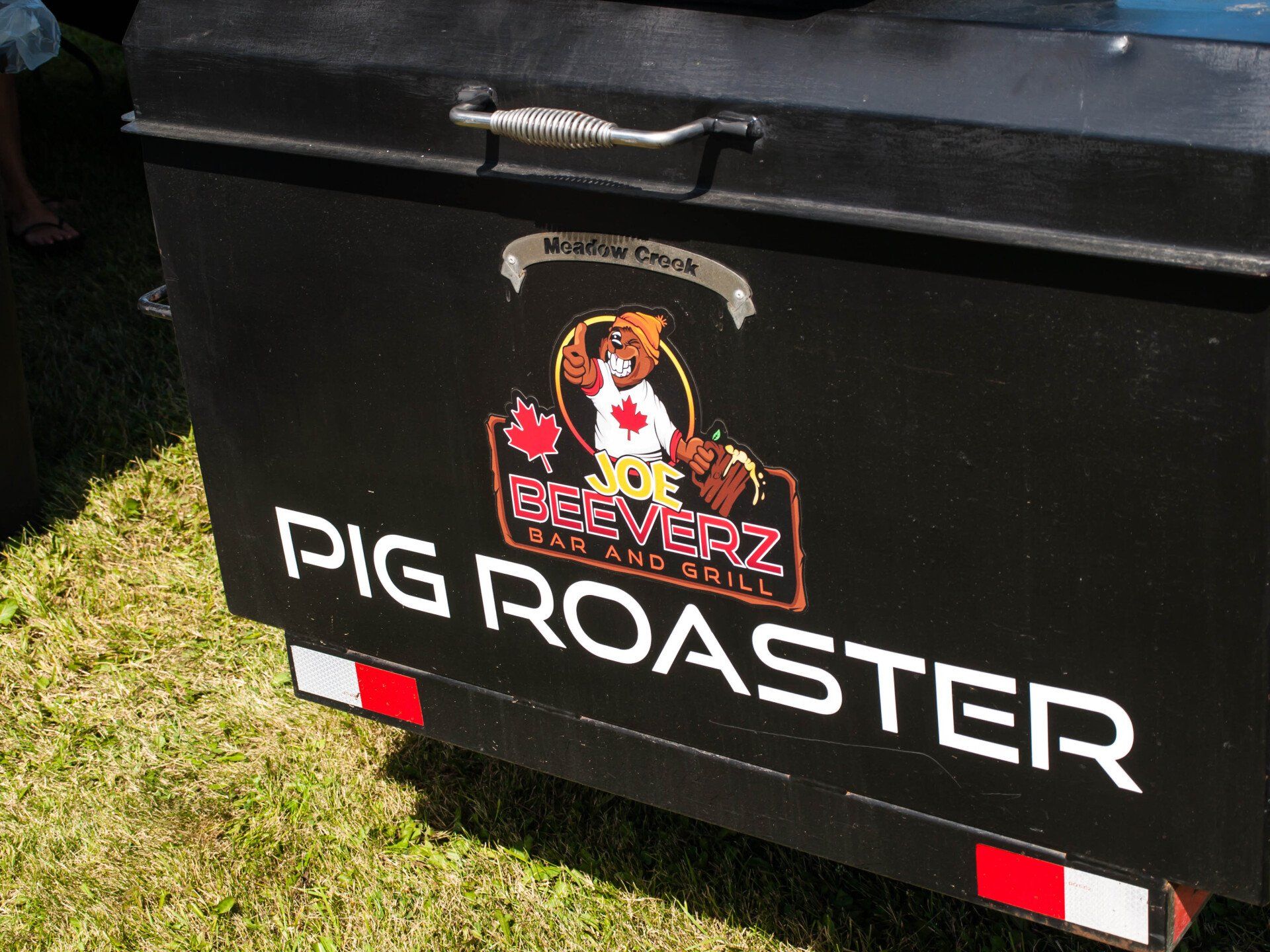 Black trailer with a pig roast logo and large white text reading “PIG ROASTER” on grass