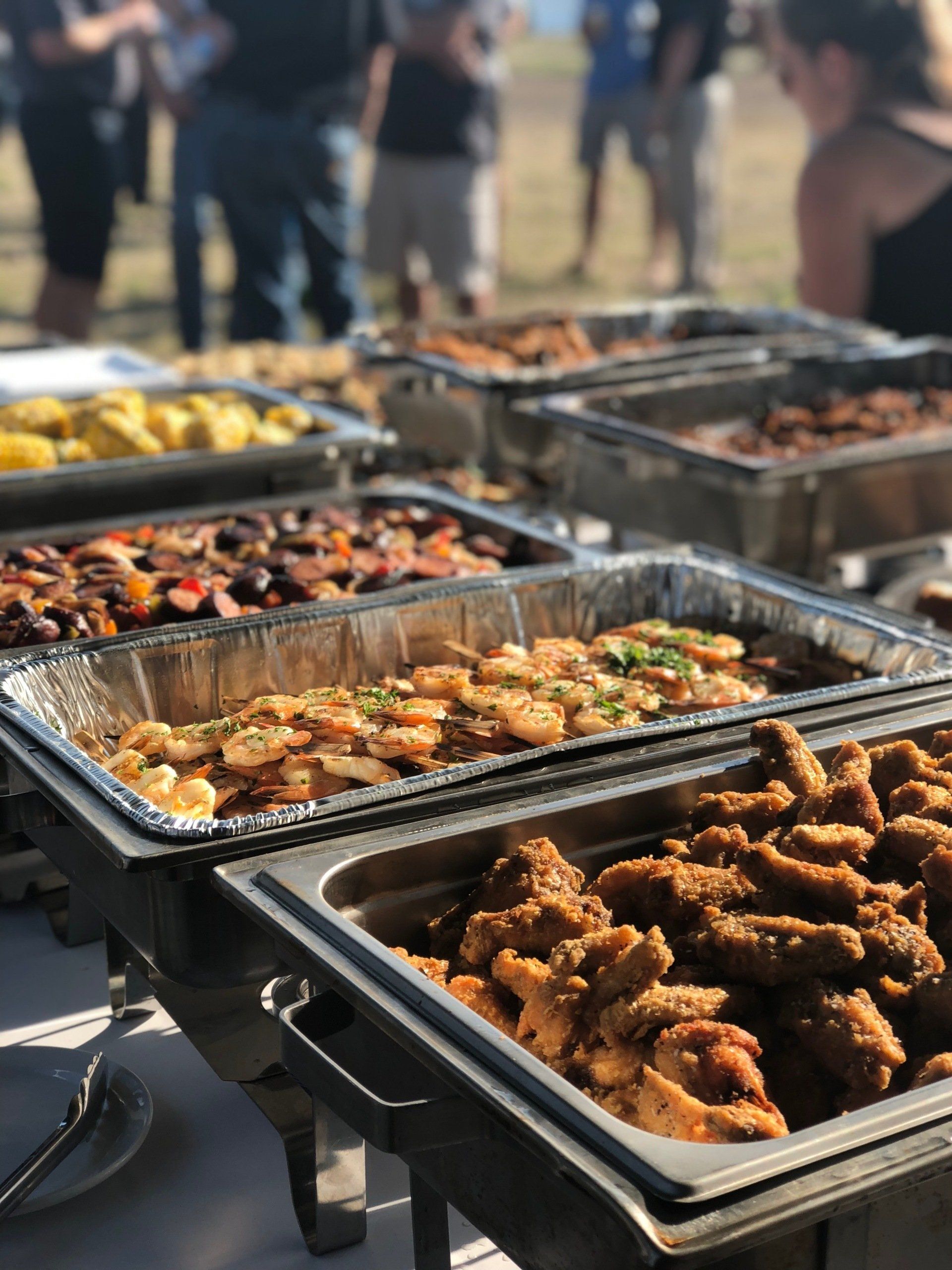 Trays of assorted grilled food at an outdoor buffet with people in the background