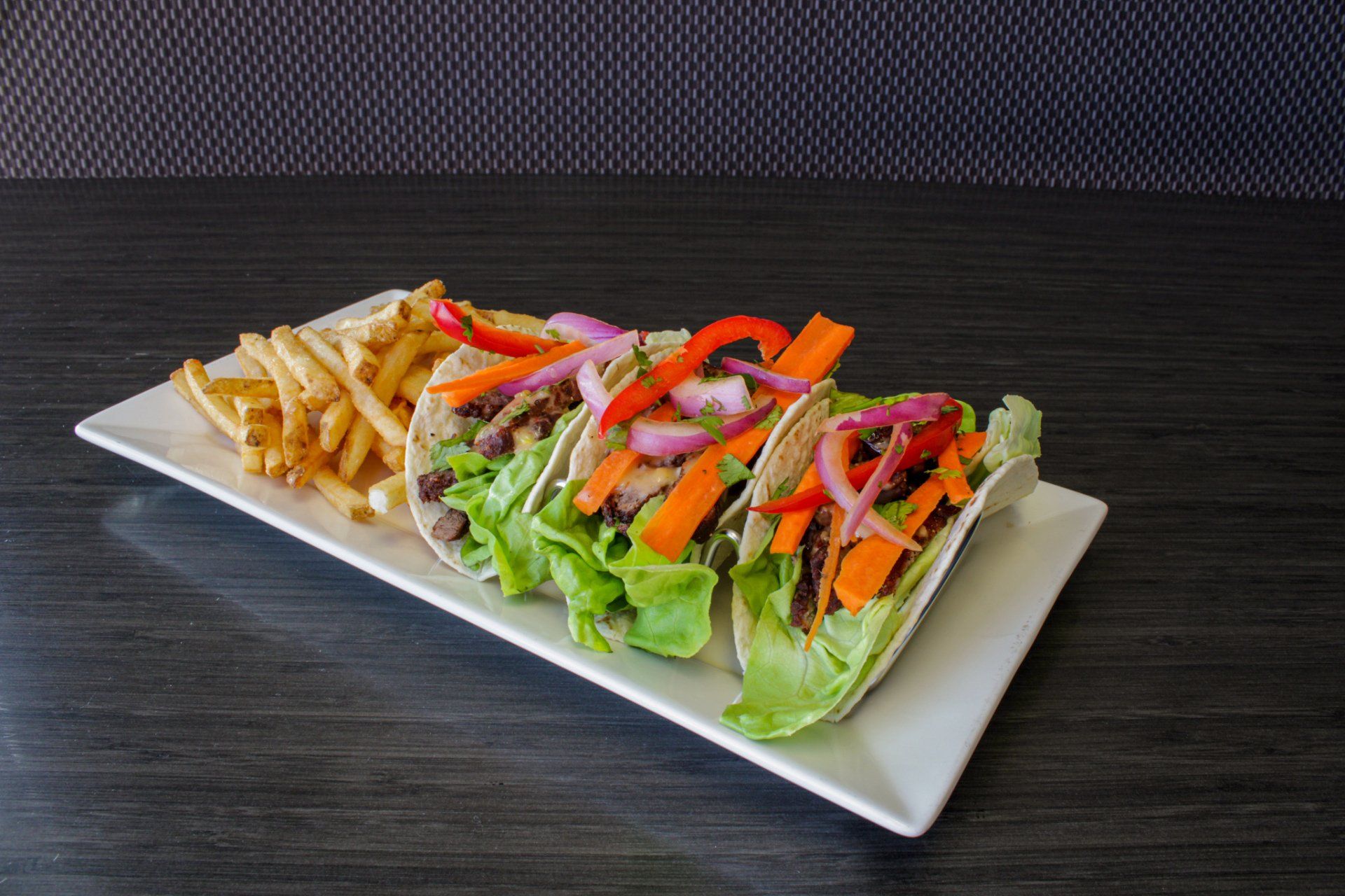 Fresh tacos with lettuce, tomatoes, and peppers on a white plate beside fries