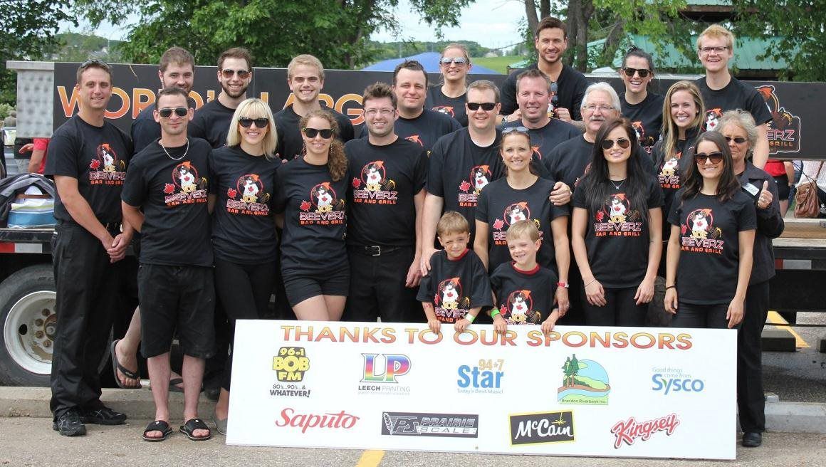 Group of people in black shirts posing outdoors behind a “Thanks to our sponsors” sign.