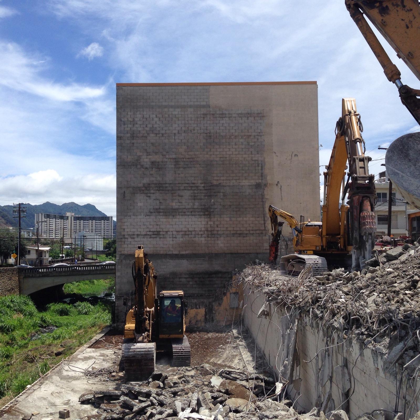 Construction equipment demolishes a concrete building next to a river. Sunny day.