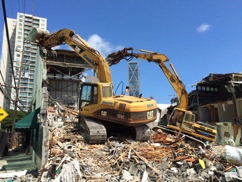 Two yellow excavators demolish a building, debris piled below, blue sky, city in background.