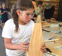 A young girl wearing safety glasses is holding a piece of wood