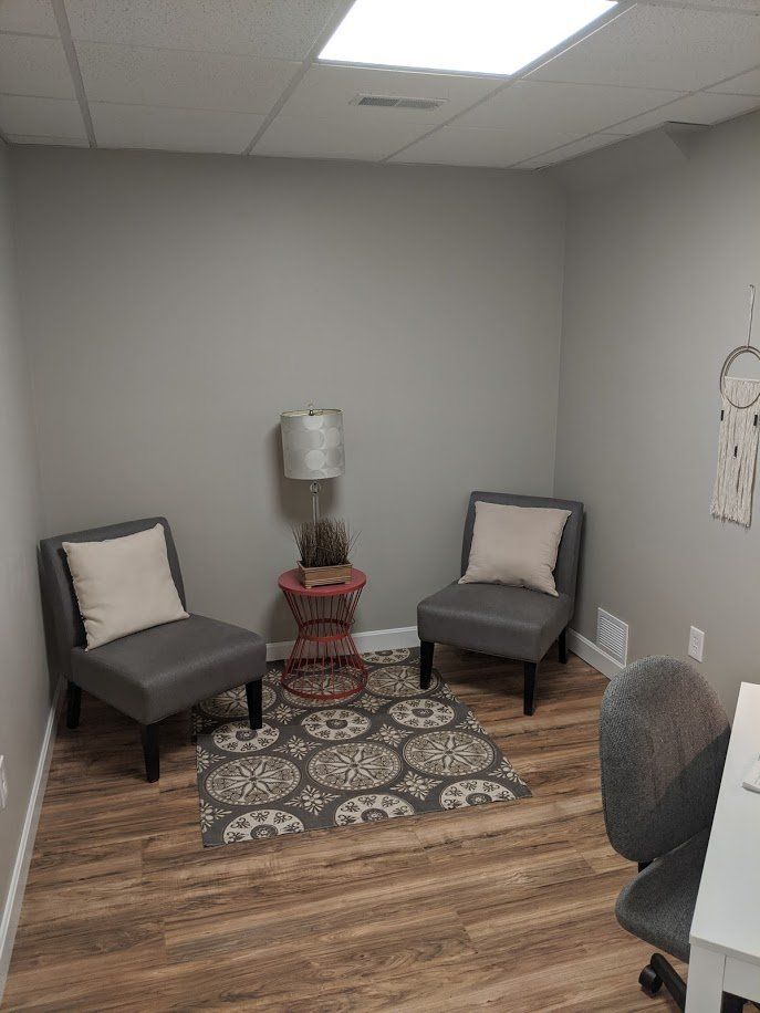 Small gray waiting nook with two armchairs, a side table, lamp, and patterned rug on wood flooring.