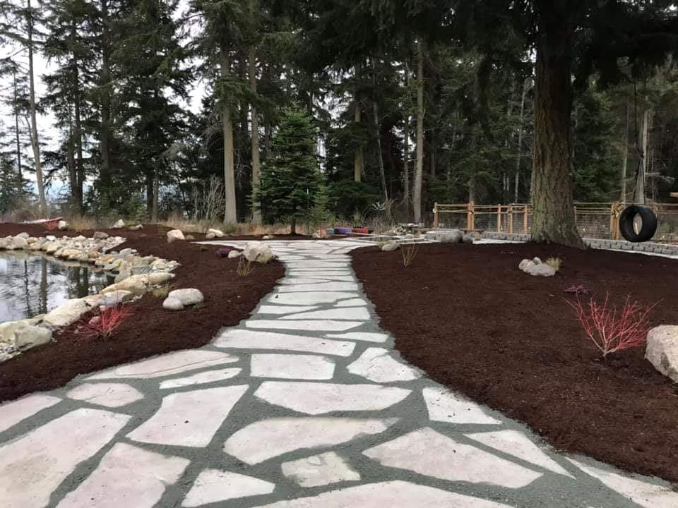 Stone path through a landscaped yard with a pond, trees, and dark mulch.