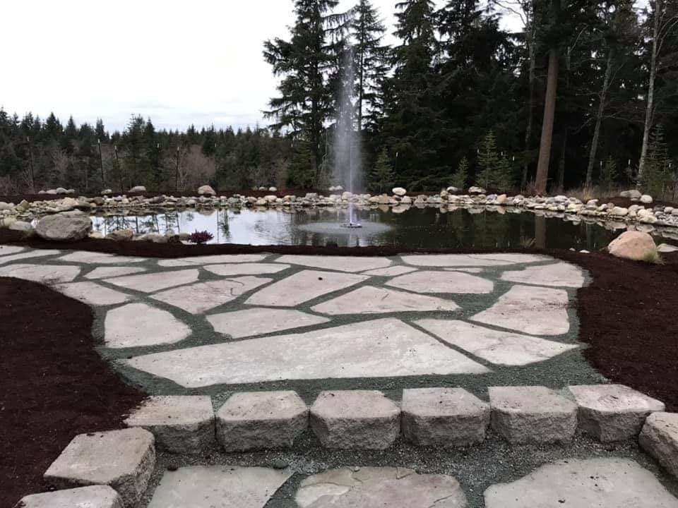 Stone patio leading to a pond with fountain, surrounded by trees and mulch.